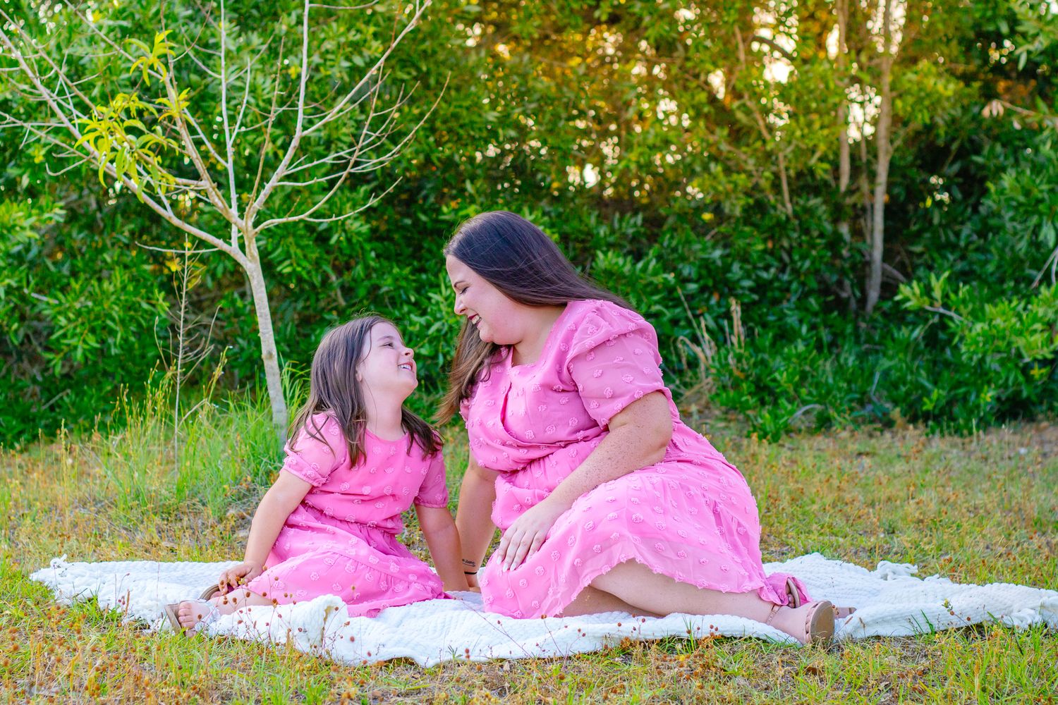A mother and daughter in matching pink dresses sit on a blanket outdoors, smiling at each other surrounded by greenery.