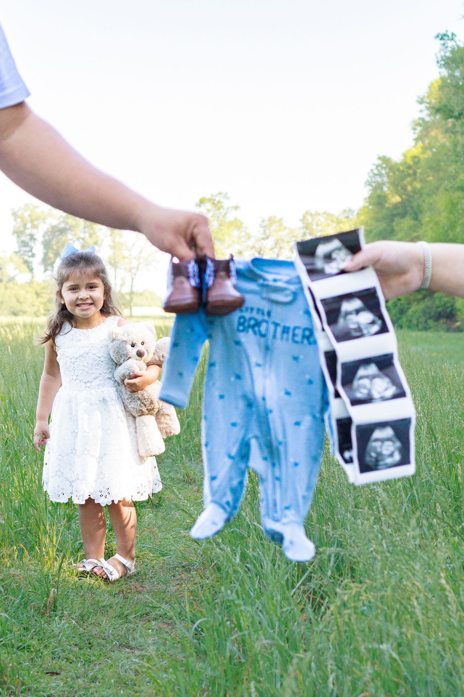 Young girl in white dress holds teddy bear as parents display Little Brother onesie, shoes, and ultrasound photos outdoors.