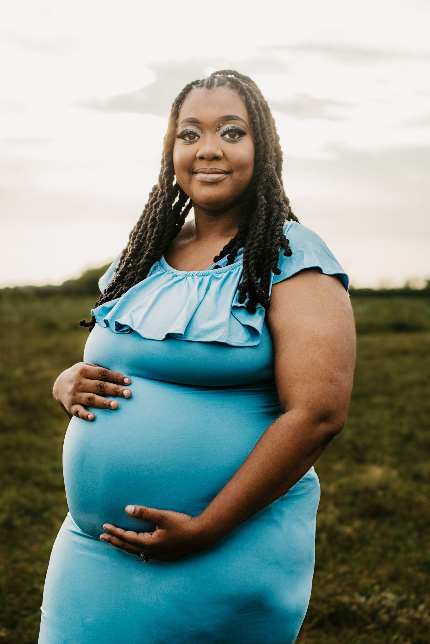 Pregnant Black woman in blue ruffle dress cradling baby bump outdoors at golden hour.