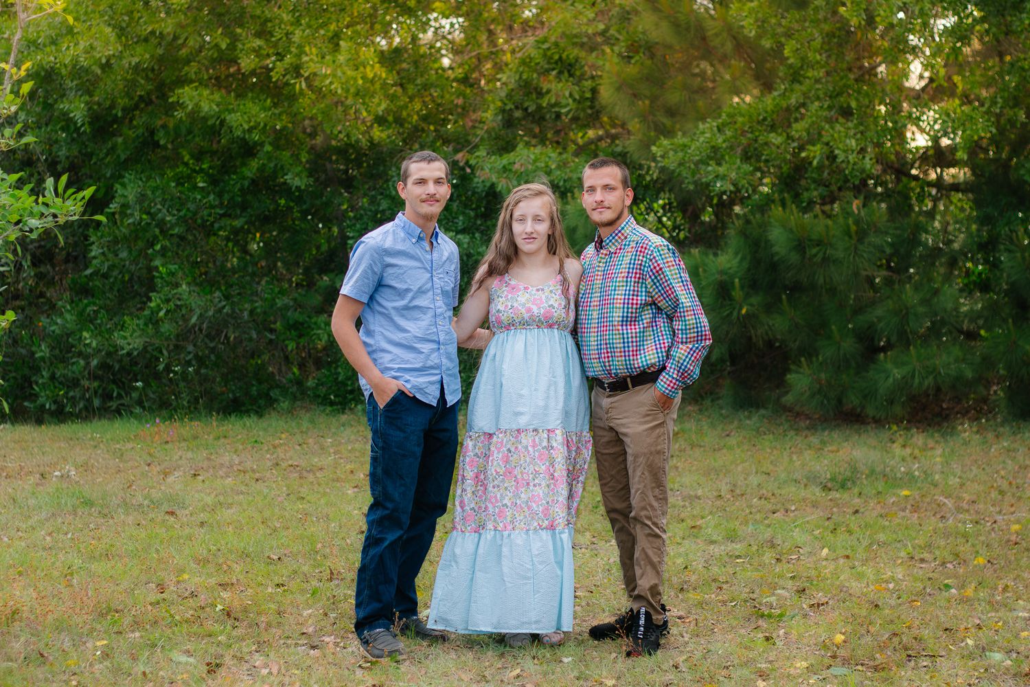 Three people posing outdoors in a grassy area with green trees, two men flanking a woman in a floral dress.