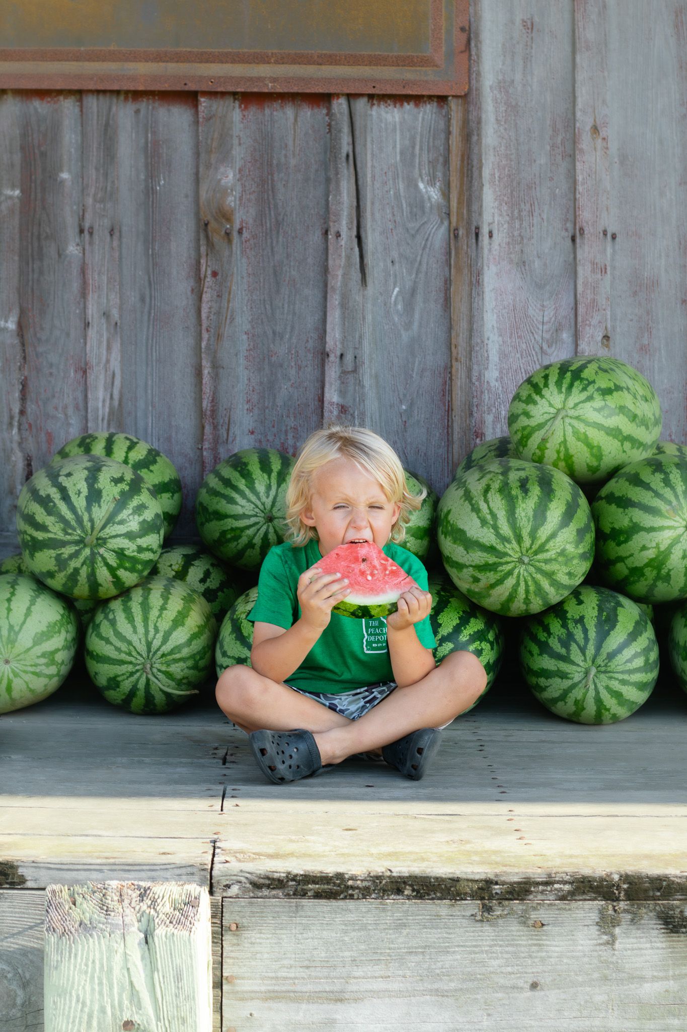 Blond child sitting cross-legged eating watermelon slice surrounded by stacked whole watermelons against rustic wooden barn.
