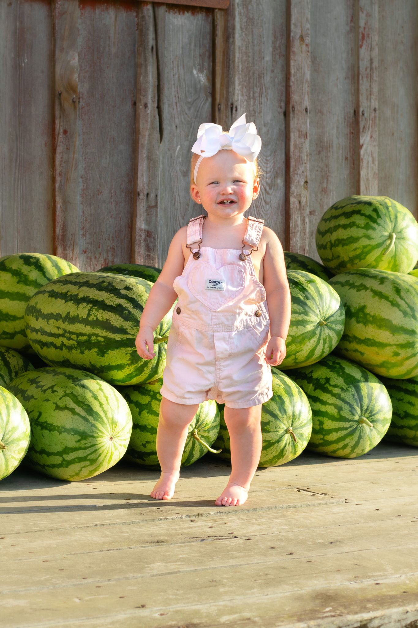 Happy baby girl in pink overalls and white hair bow stands smiling in front of stacked watermelons outdoors.