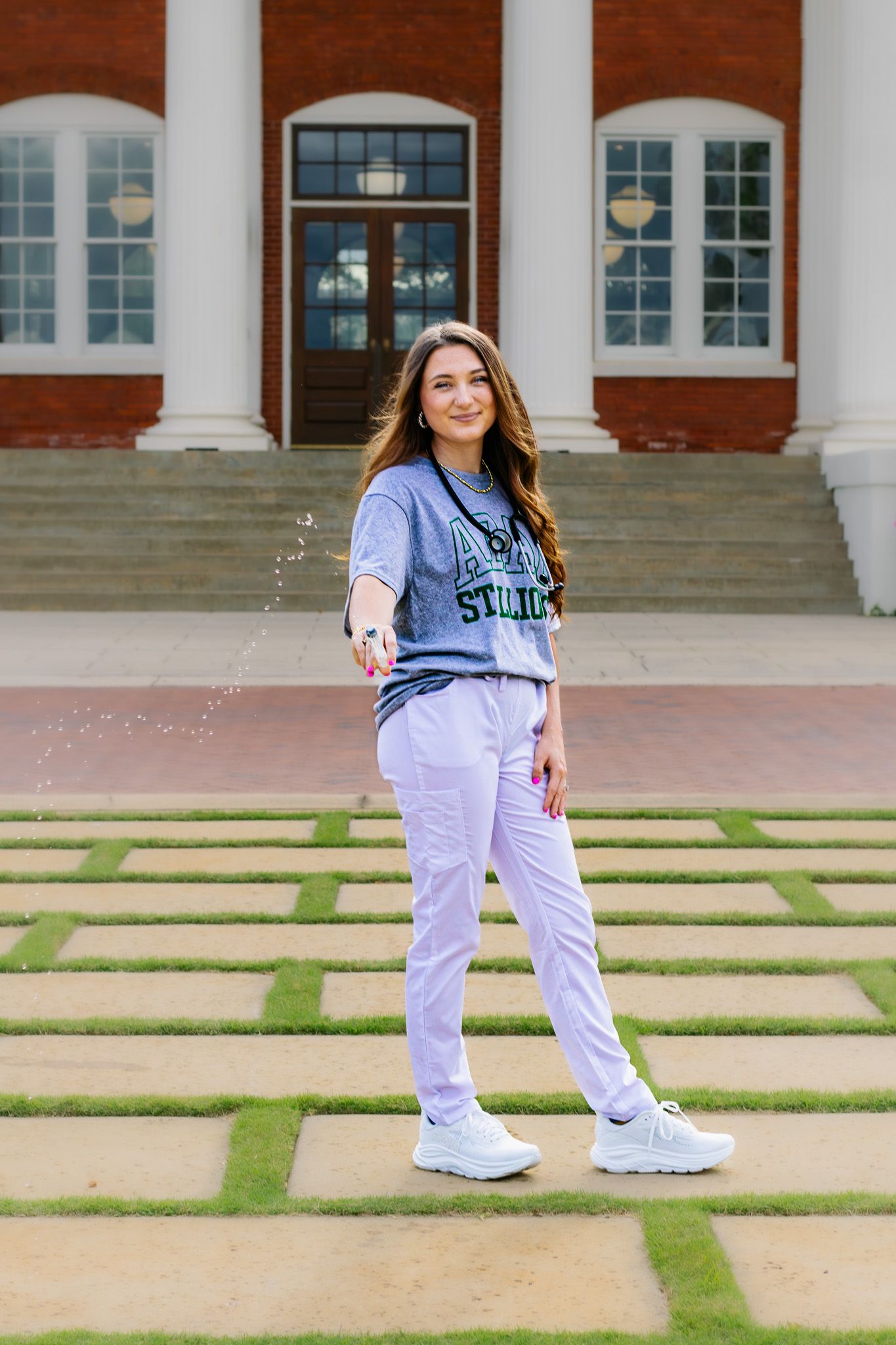 Smiling young woman in gray college shirt and white pants tossing water outside a brick building.