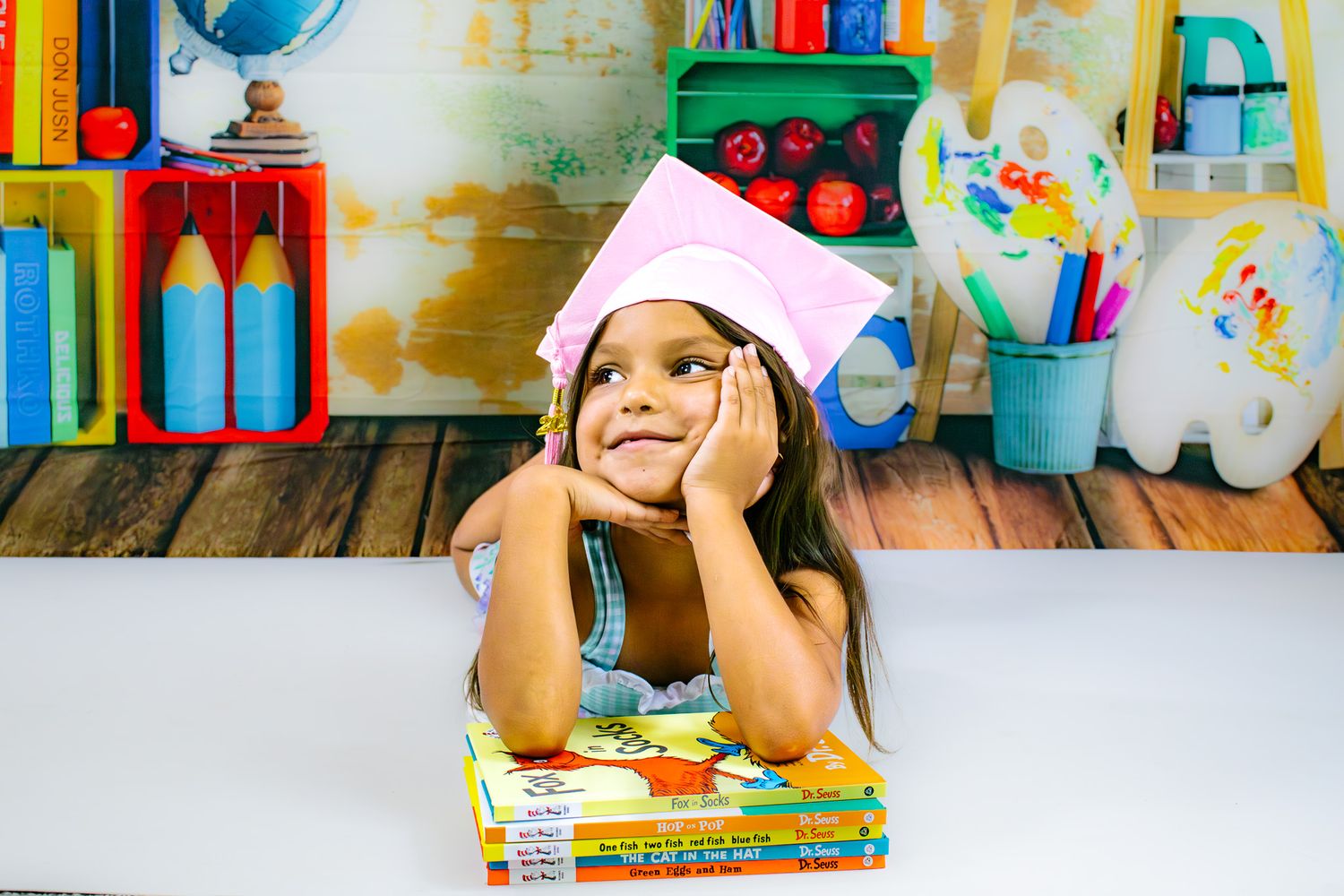 Young girl wearing a pink graduation cap rests her chin on hands, sitting with colorful children's books in a bright classroom.