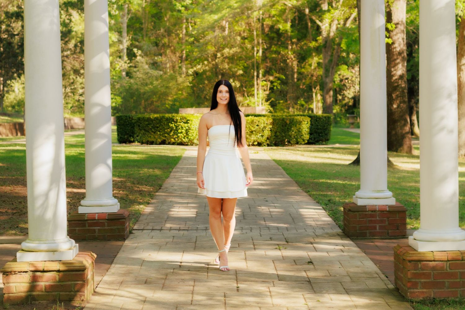 Young woman in white dress walking between white columns on brick path in lush green garden setting.