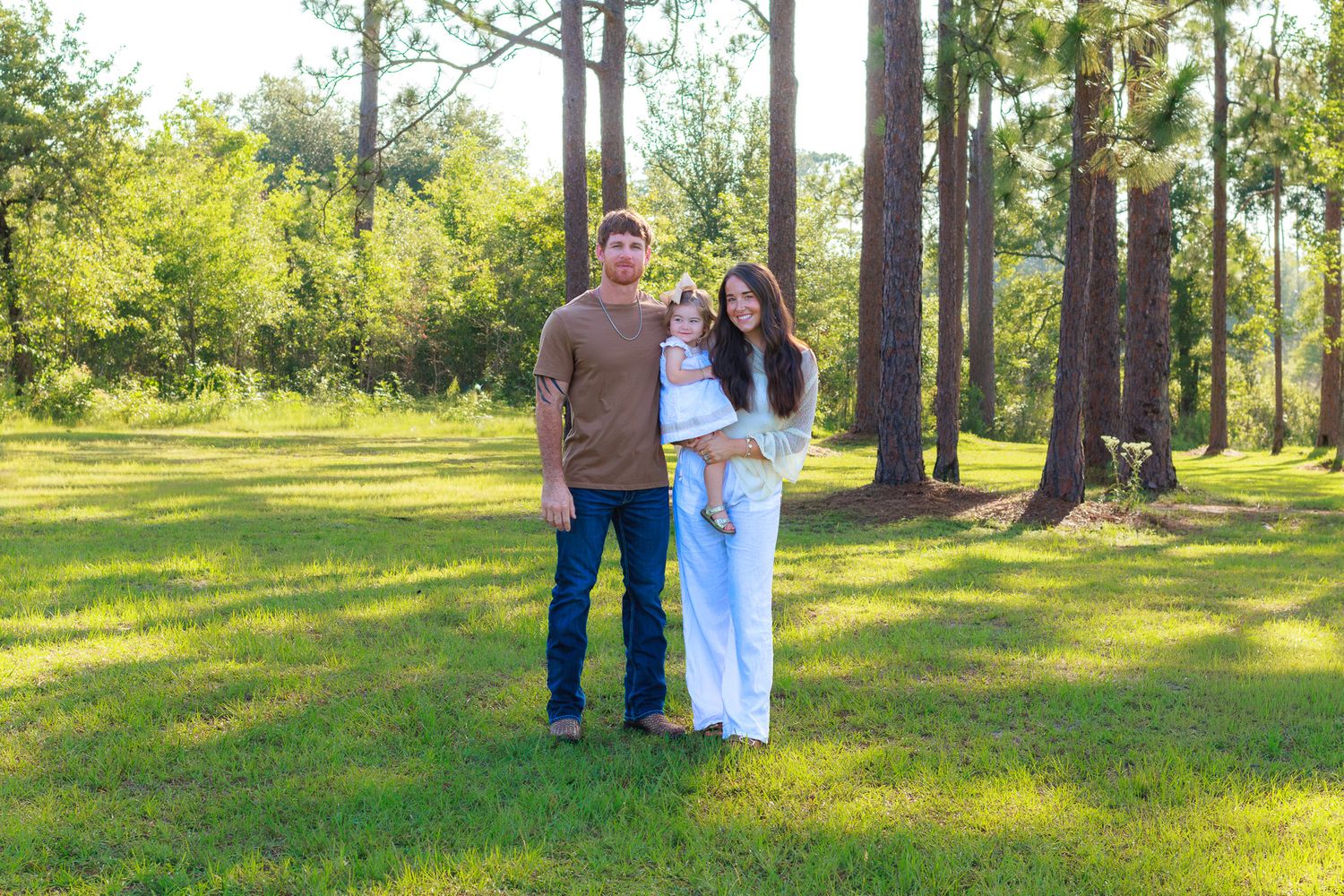 Young family with baby posing outdoors on green grass surrounded by tall pine trees on a sunny day.
