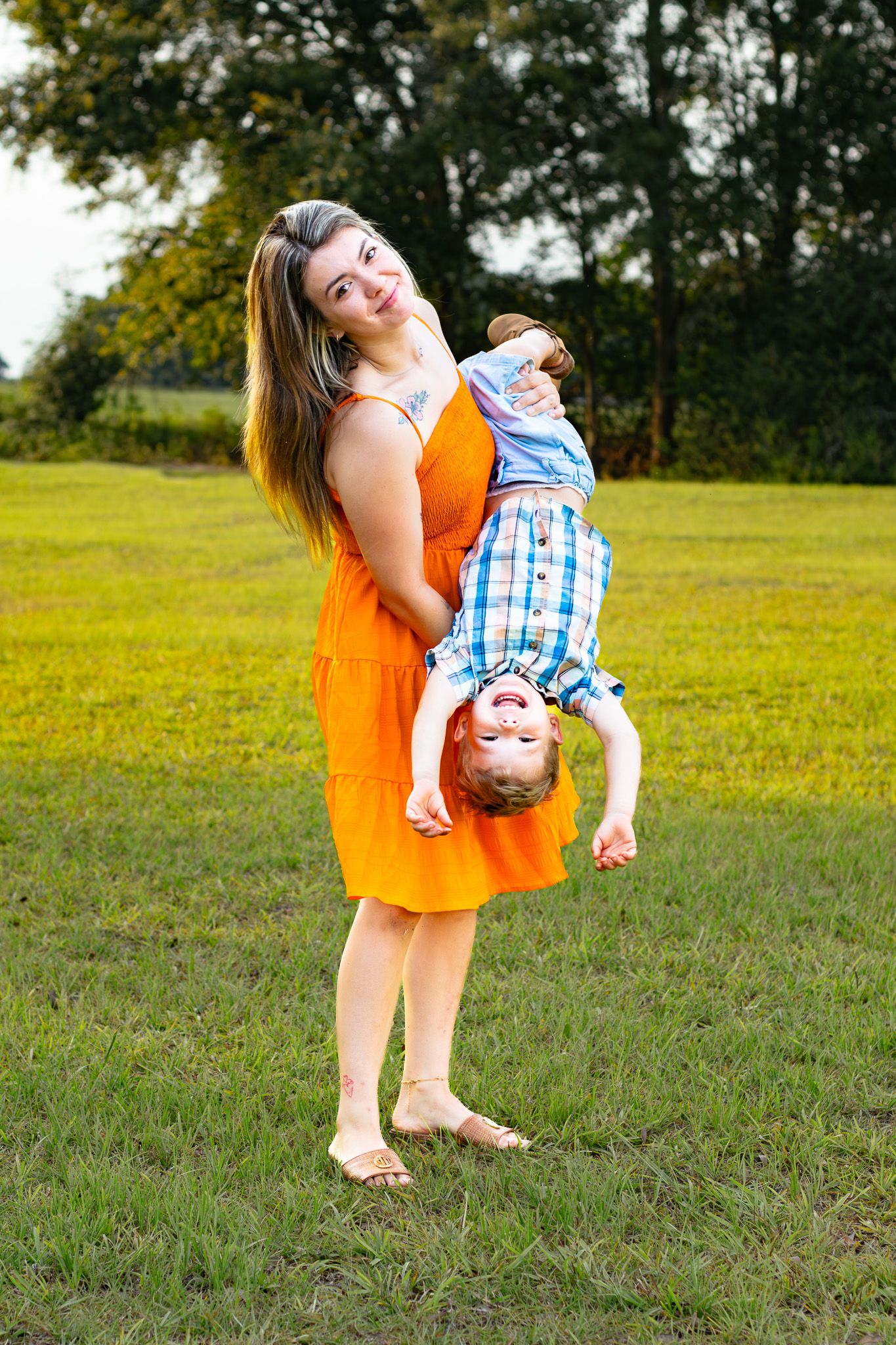 A mother in an orange dress playfully holds her young son upside down while standing barefoot on green grass outdoors.