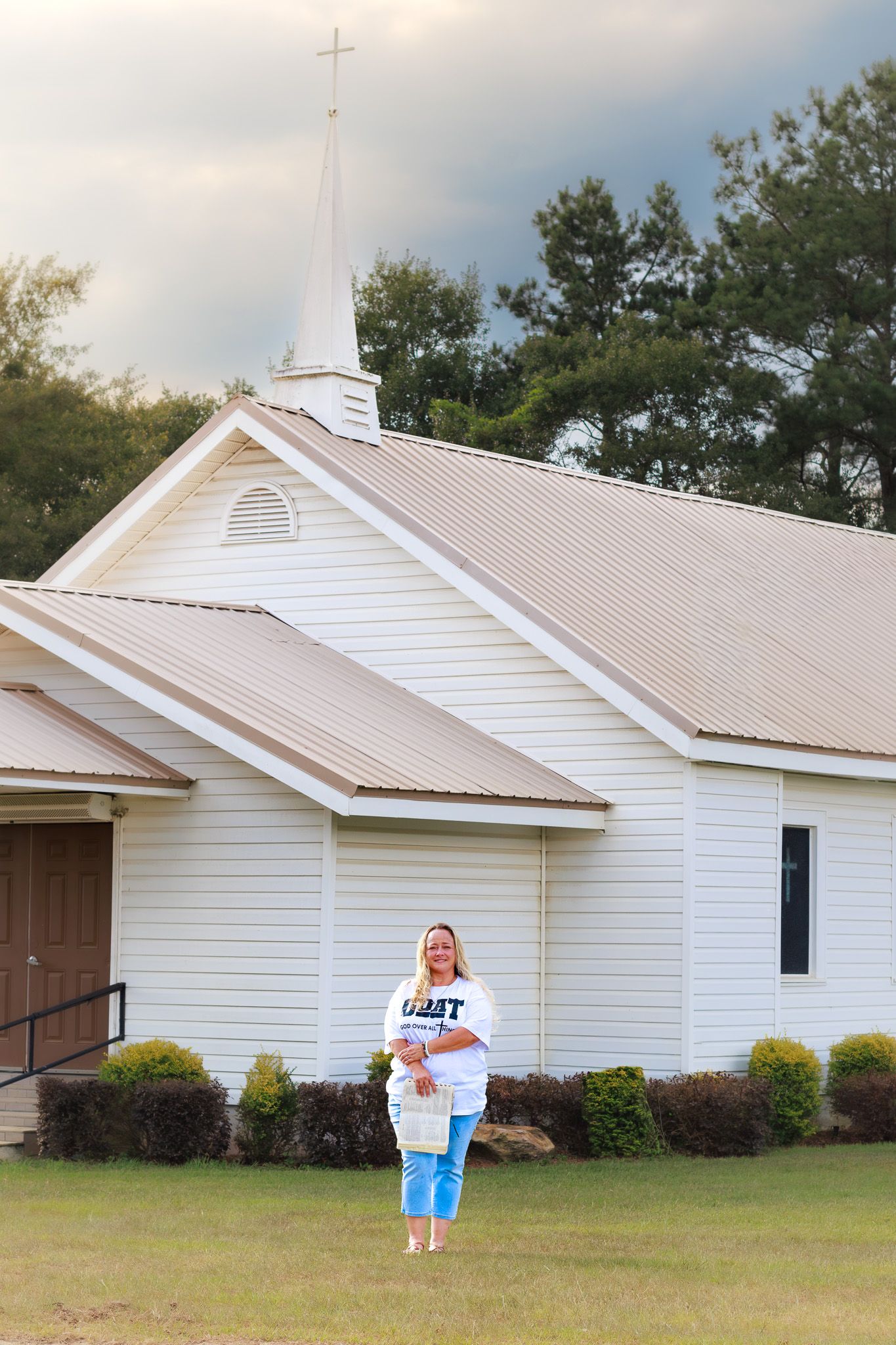 Woman standing barefoot on grass in front of a white wooden church with a steeple cross under dramatic skies.