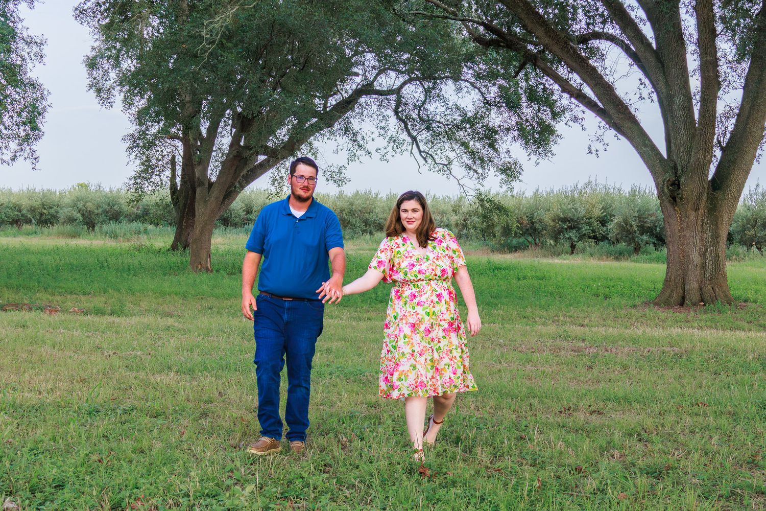 Couple holding hands walking in a green park with large trees during an outdoor engagement photo session.
