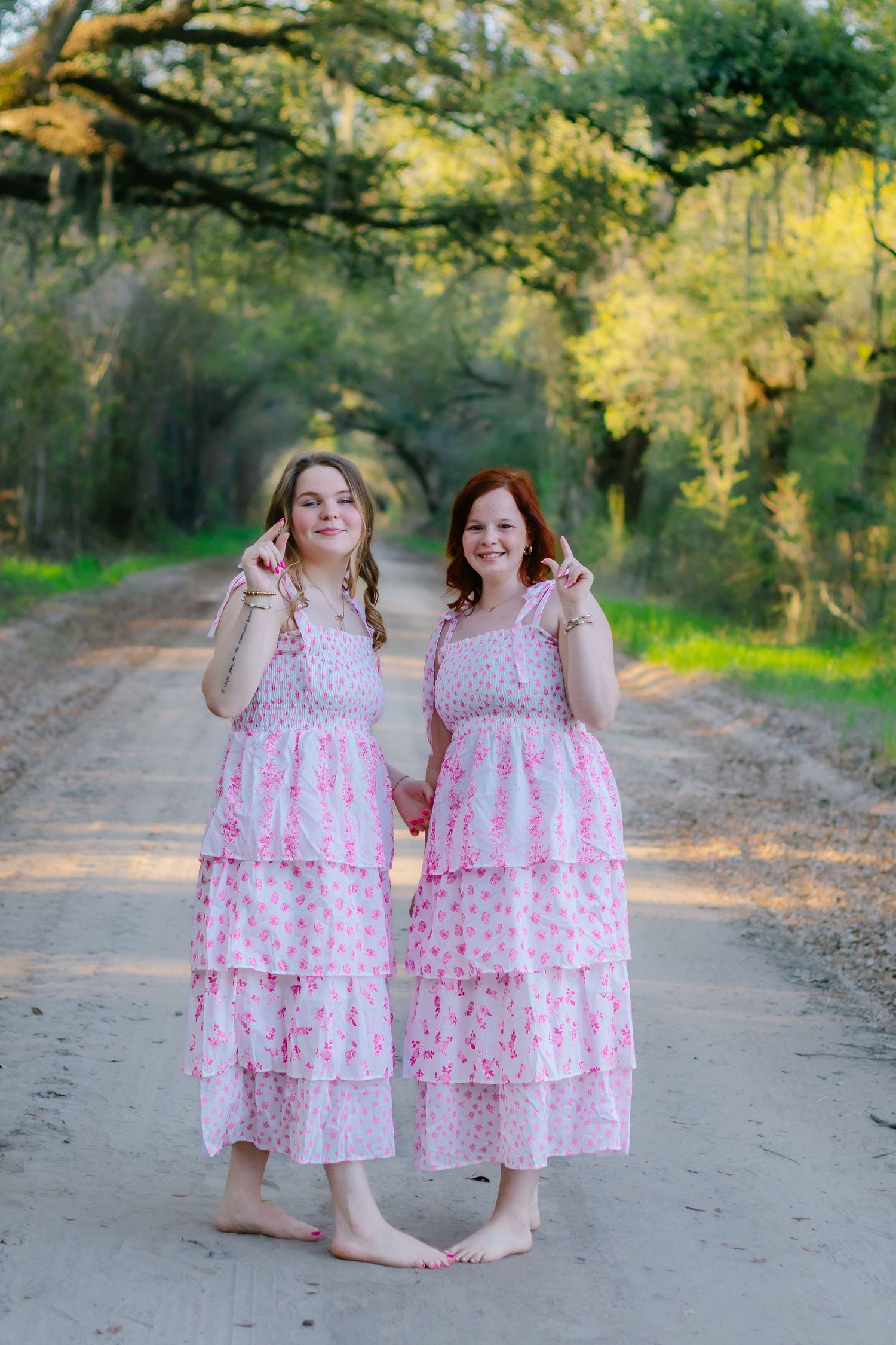 Two barefoot women in matching pink tiered dresses pose on a tree-lined dirt road giving peace signs.