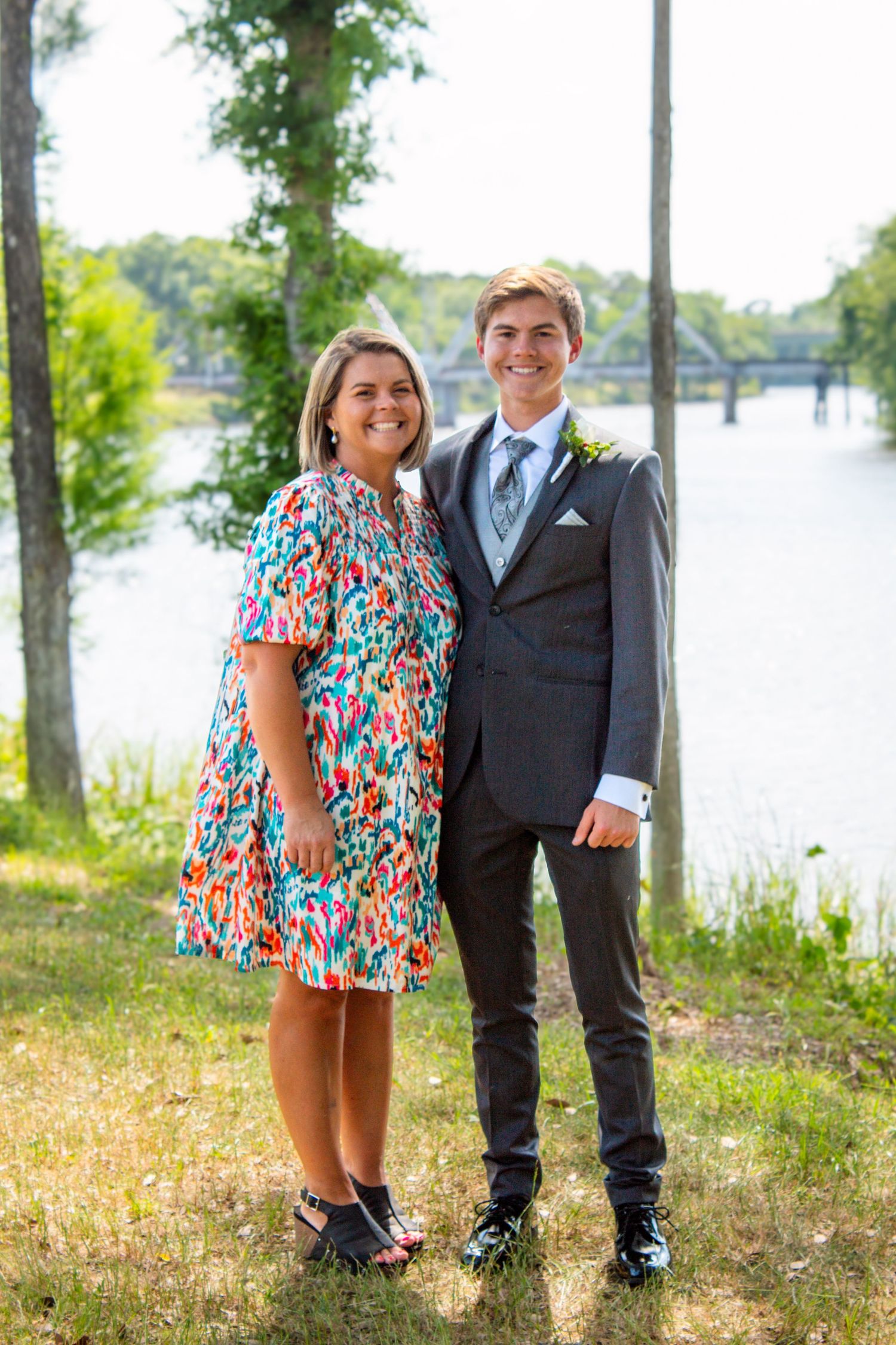 Young man in gray suit with boutonniere poses with smiling woman in colorful floral dress near a river.