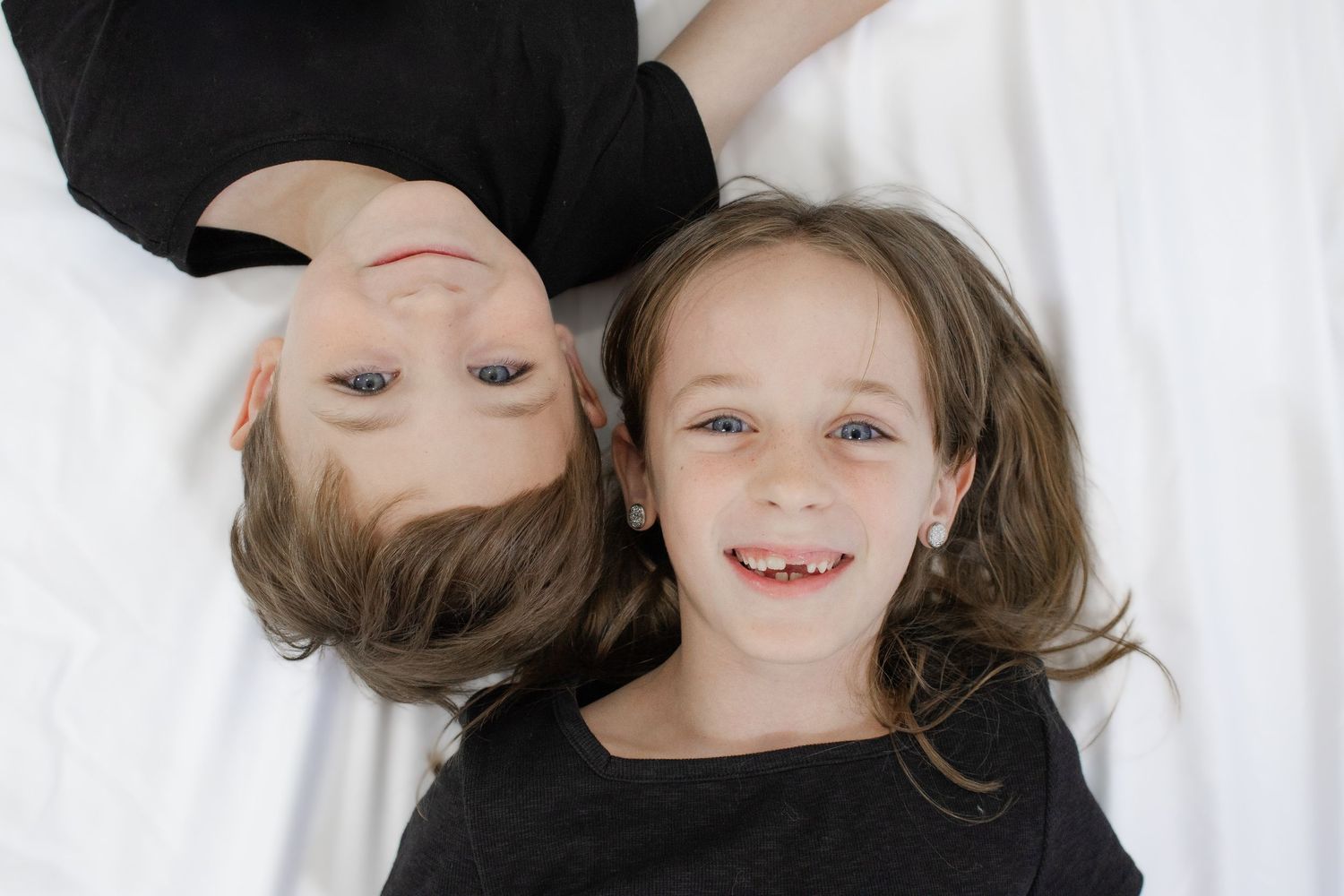 Two happy children in black shirts lying on a white background, one upside down, both smiling at the camera.
