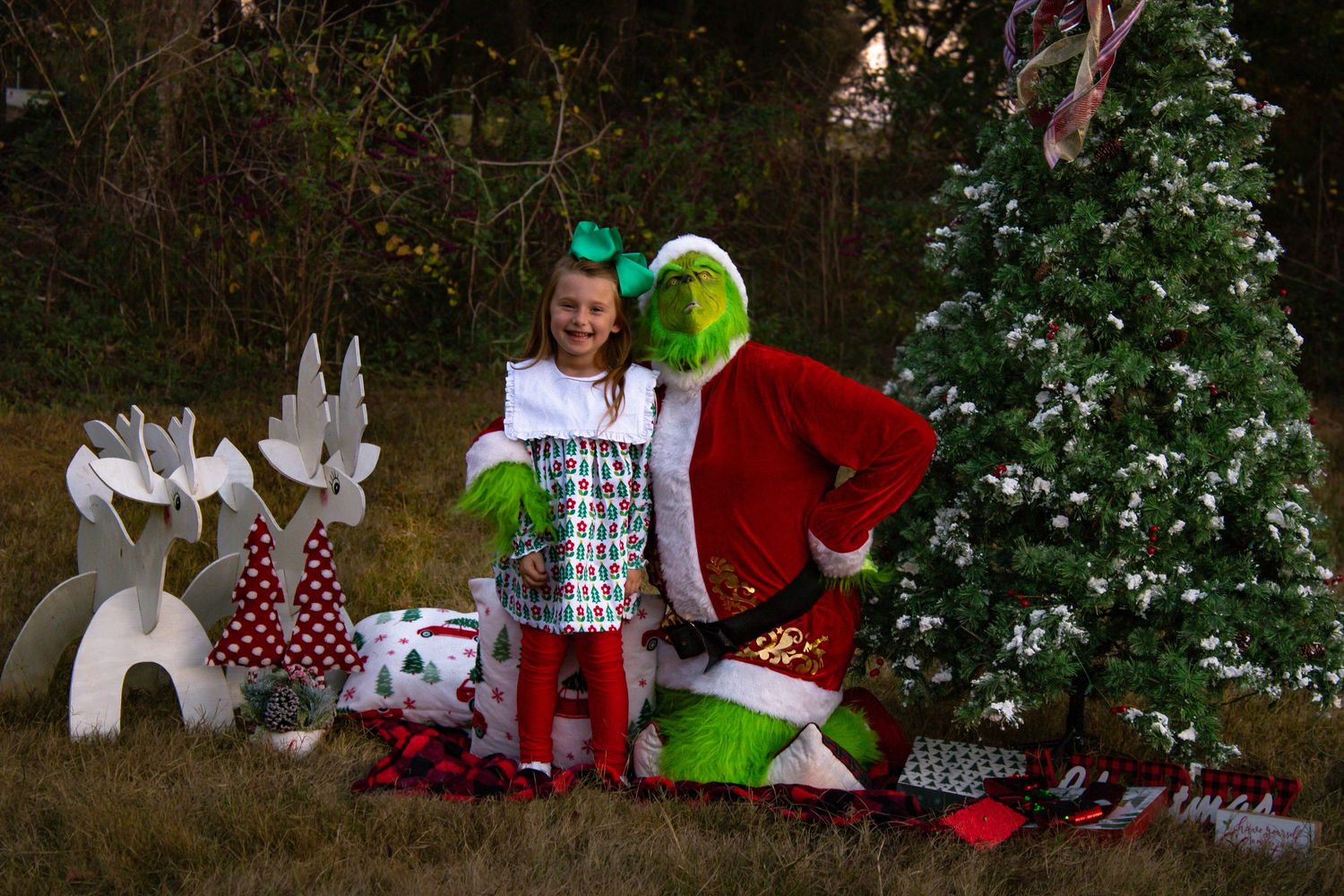 Young girl posing with Grinch character beside decorated Christmas tree and white reindeer cutouts.