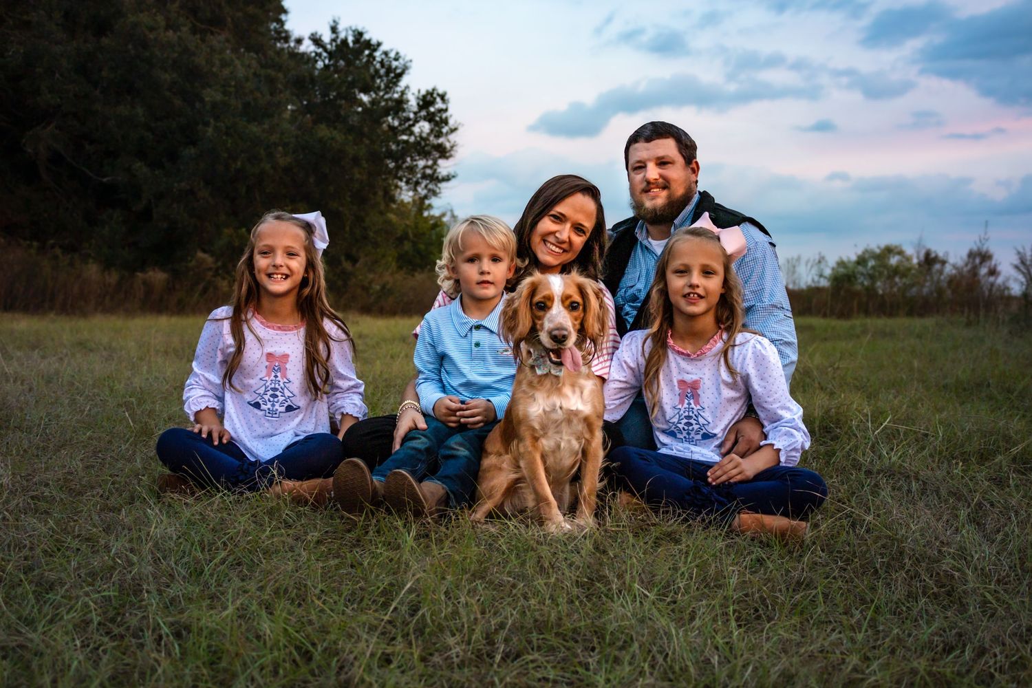 Happy family of five with a Spaniel dog sitting in a grassy field at sunset.