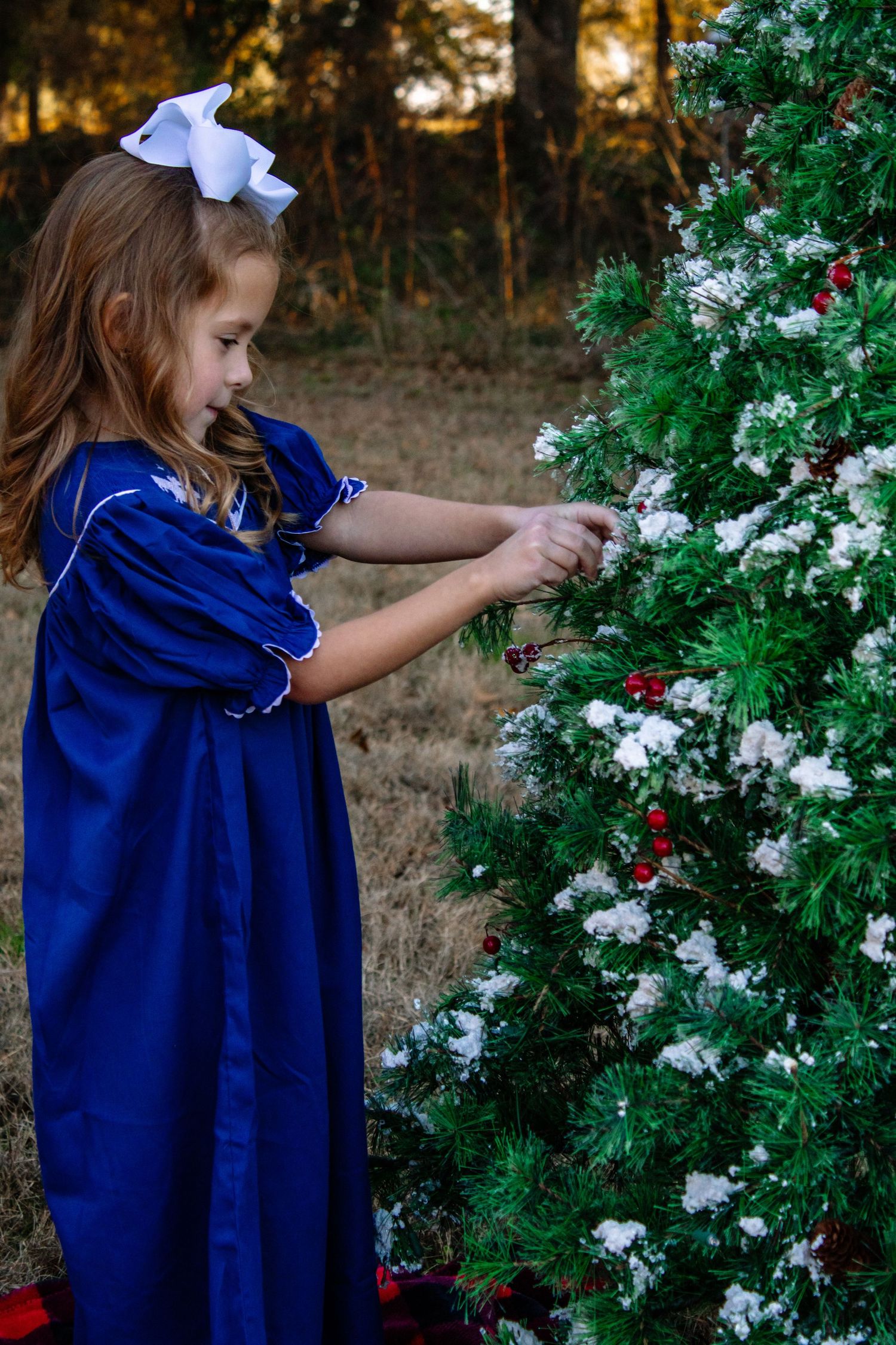 Young girl in blue dress decorates outdoor Christmas tree with white bow in hair.