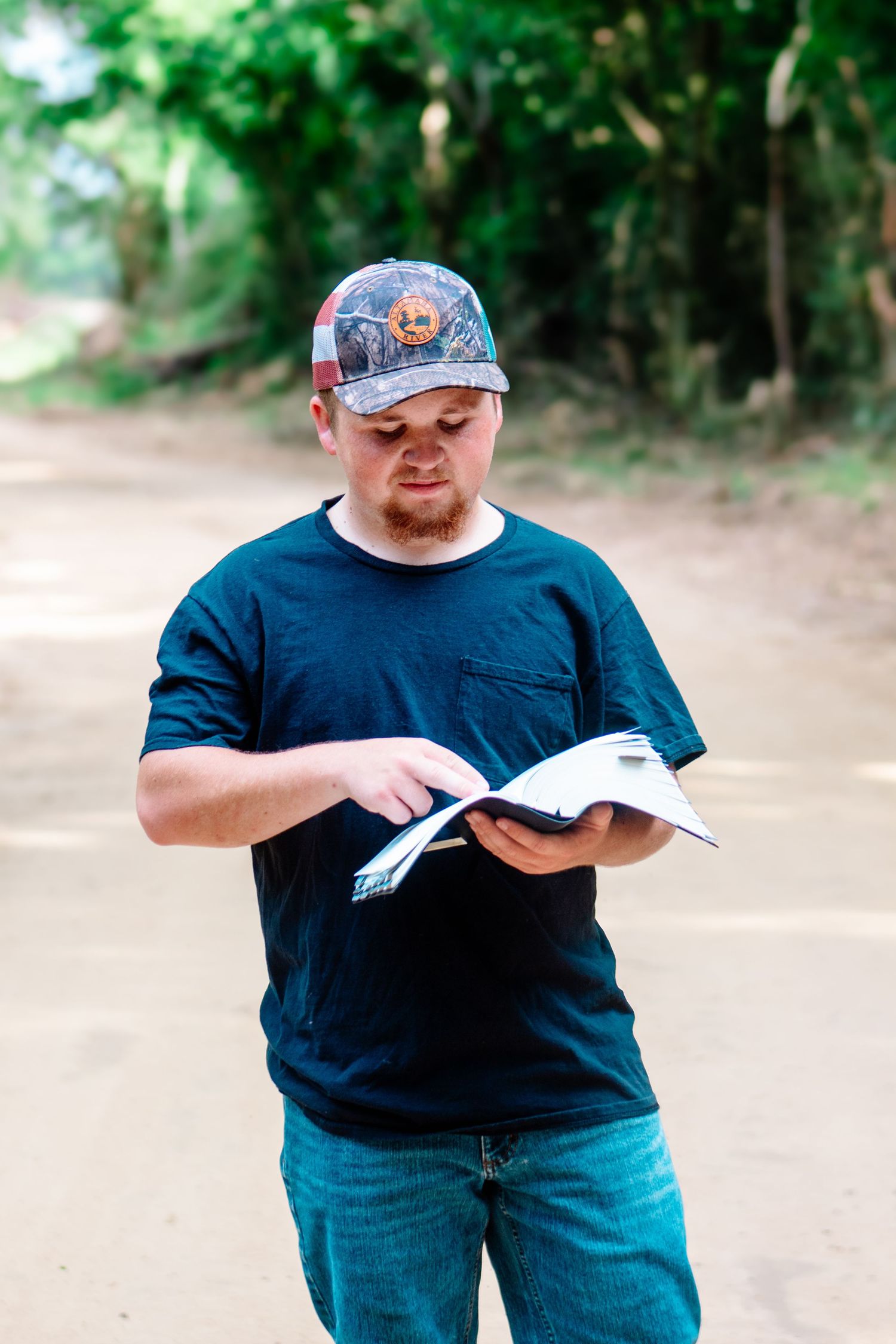 Man in black t-shirt and camo cap reading an open book while standing on a rural dirt road.