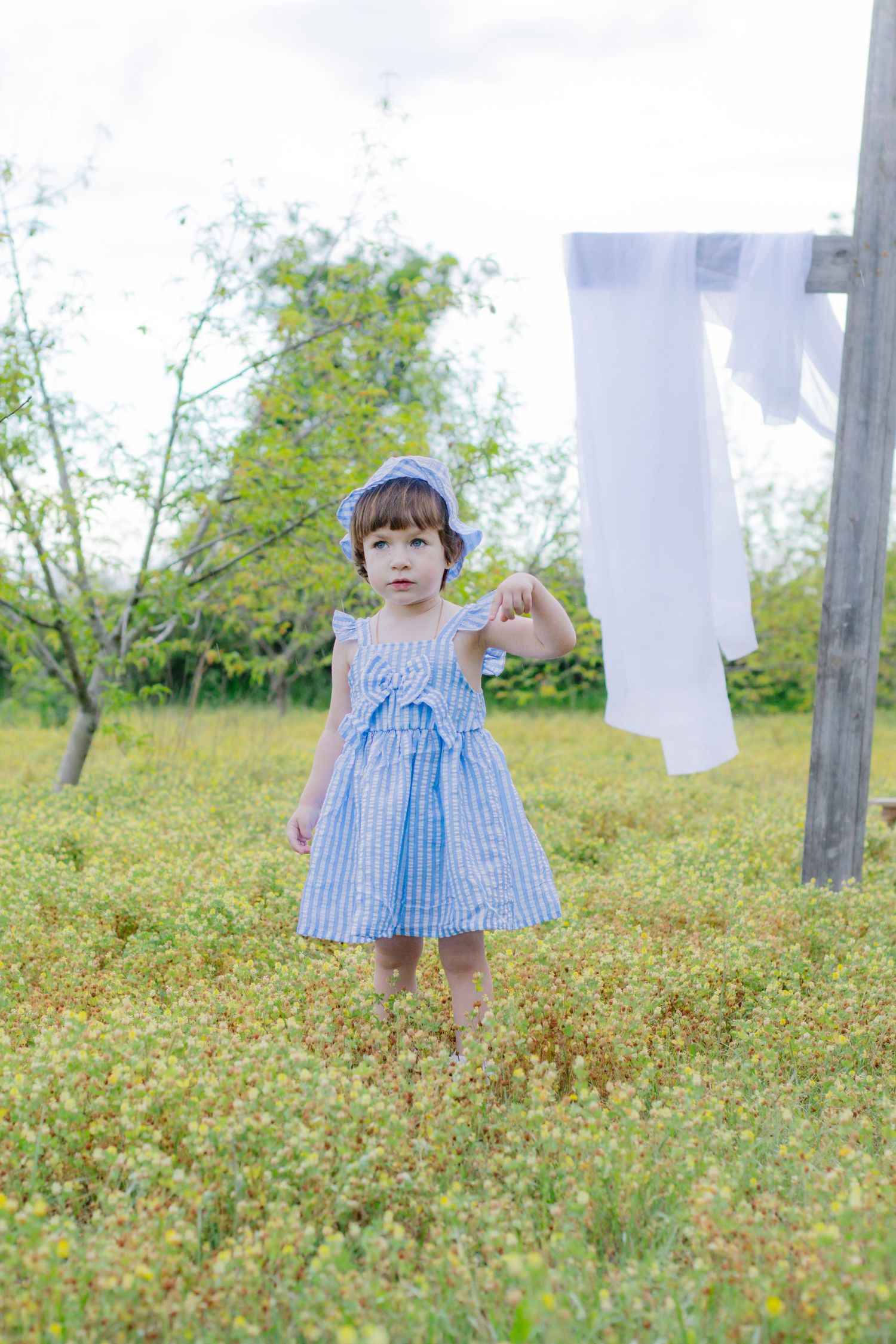 Little girl in blue striped dress and bonnet standing in green meadow near white laundry on clothesline.