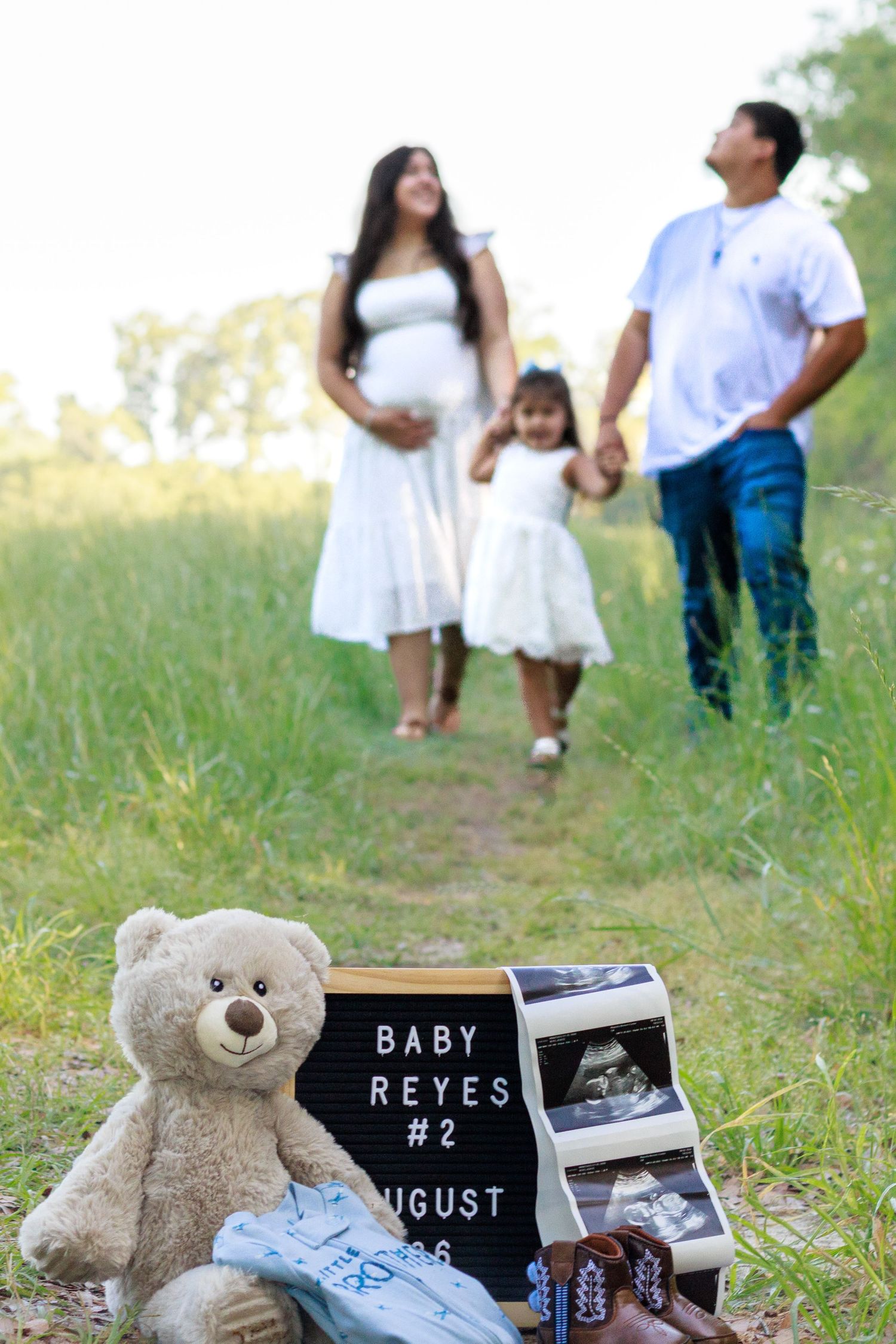 Family maternity photo with Baby Reyes #2 sign, teddy bear, ultrasound, and baby shoes in foreground.