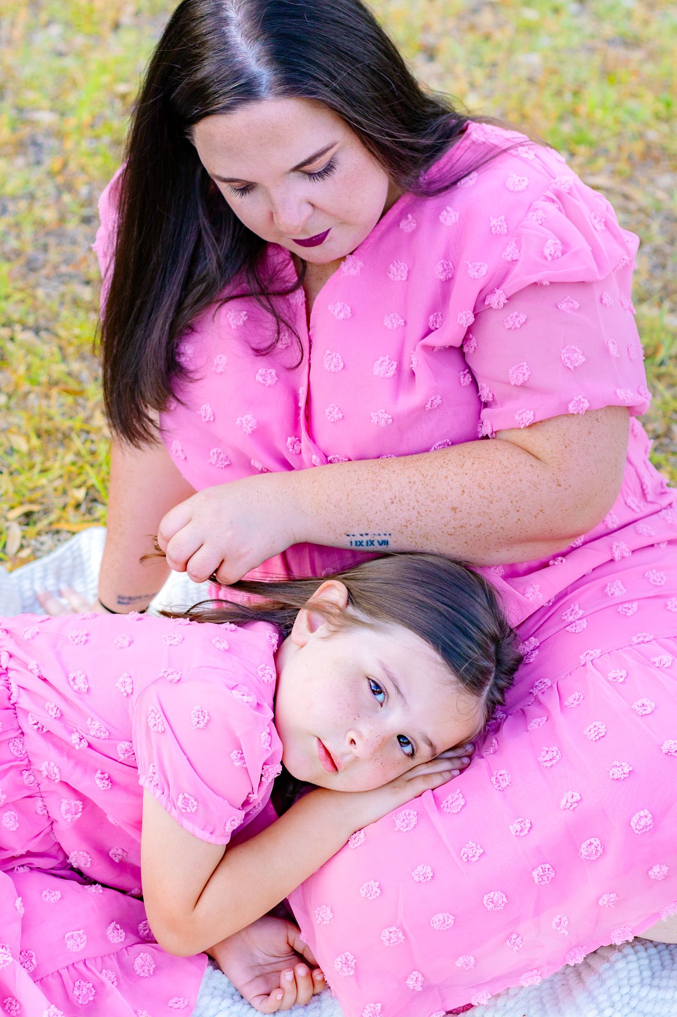A mother in a pink dress braids her daughter's hair while sitting outdoors on a blanket.