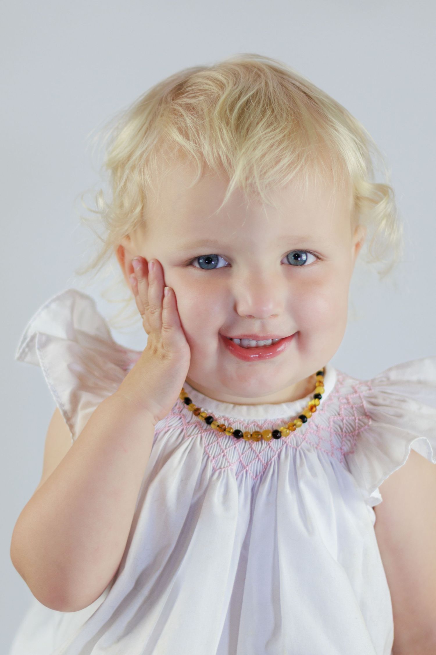 Smiling toddler girl with blonde curls wearing a white dress and amber teething necklace poses with hand on cheek.