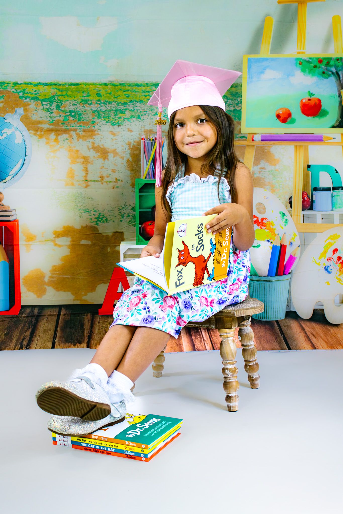 Young girl in pink graduation cap reads Dr. Seuss book, sitting in colorful classroom setting with school supplies.