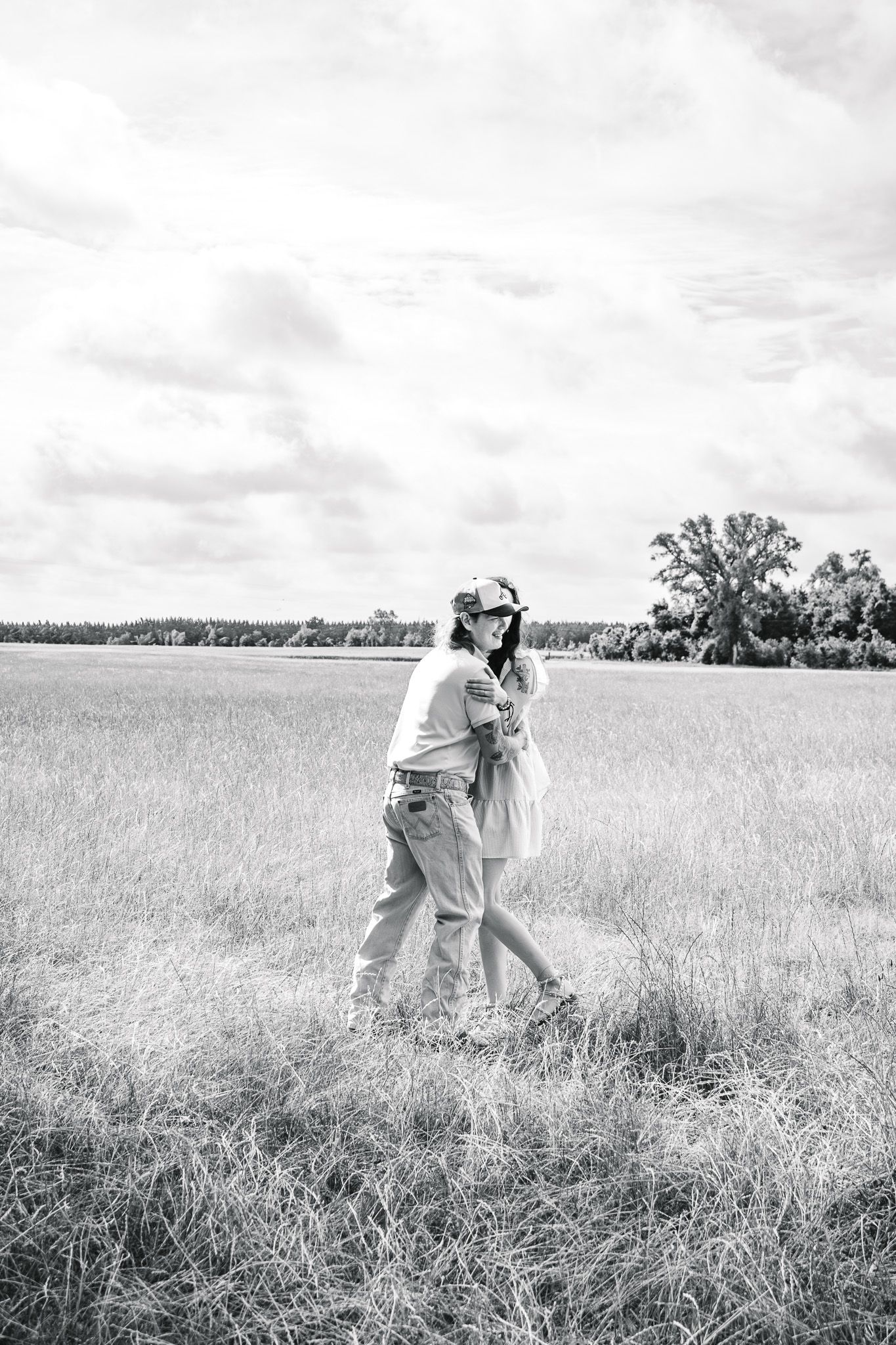 Black and white photo of a couple embracing while walking through an open grassy field under a cloudy sky.