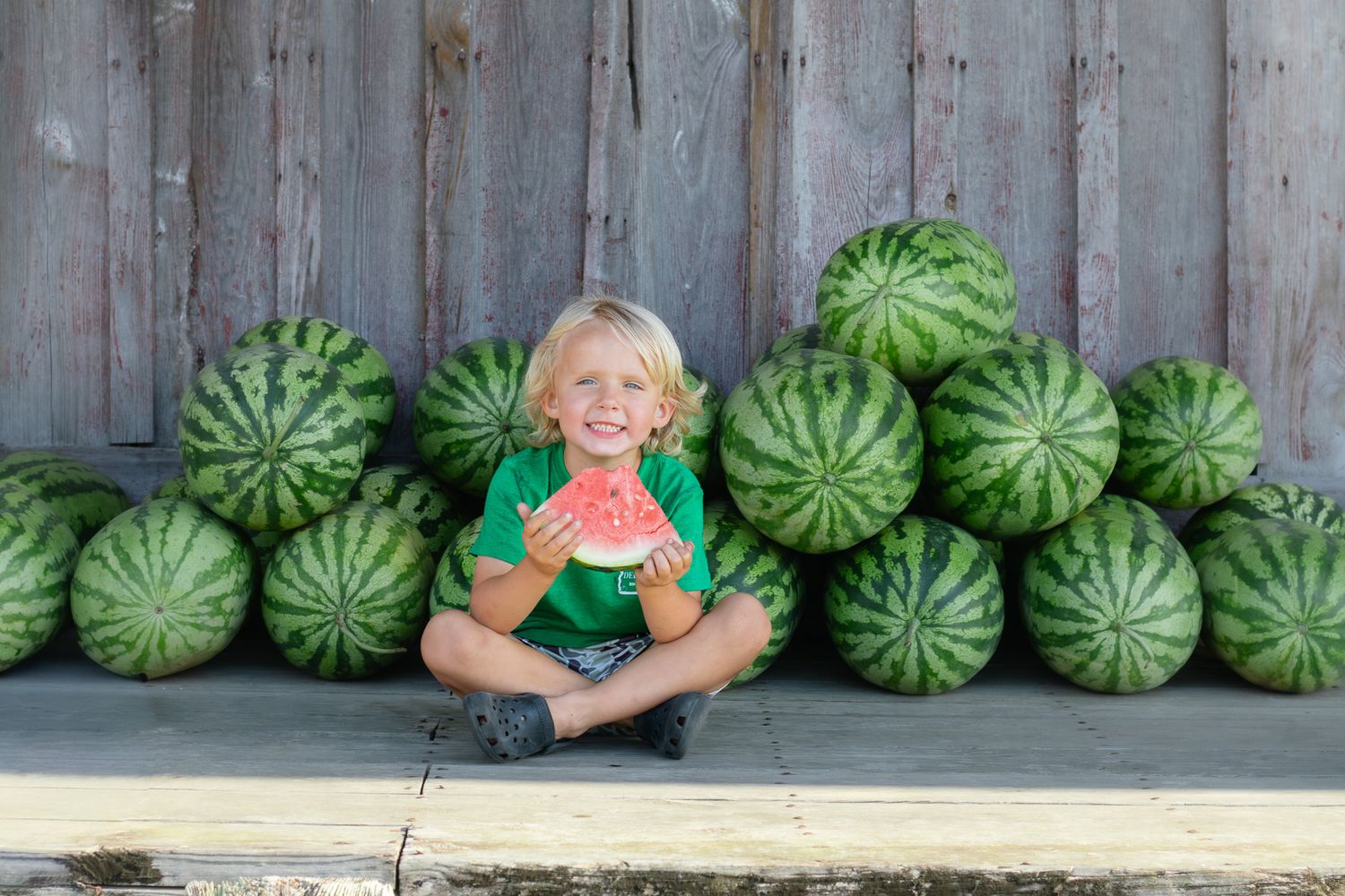 Smiling blond boy sitting cross-legged eating watermelon slice surrounded by pile of watermelons against rustic wood wall.