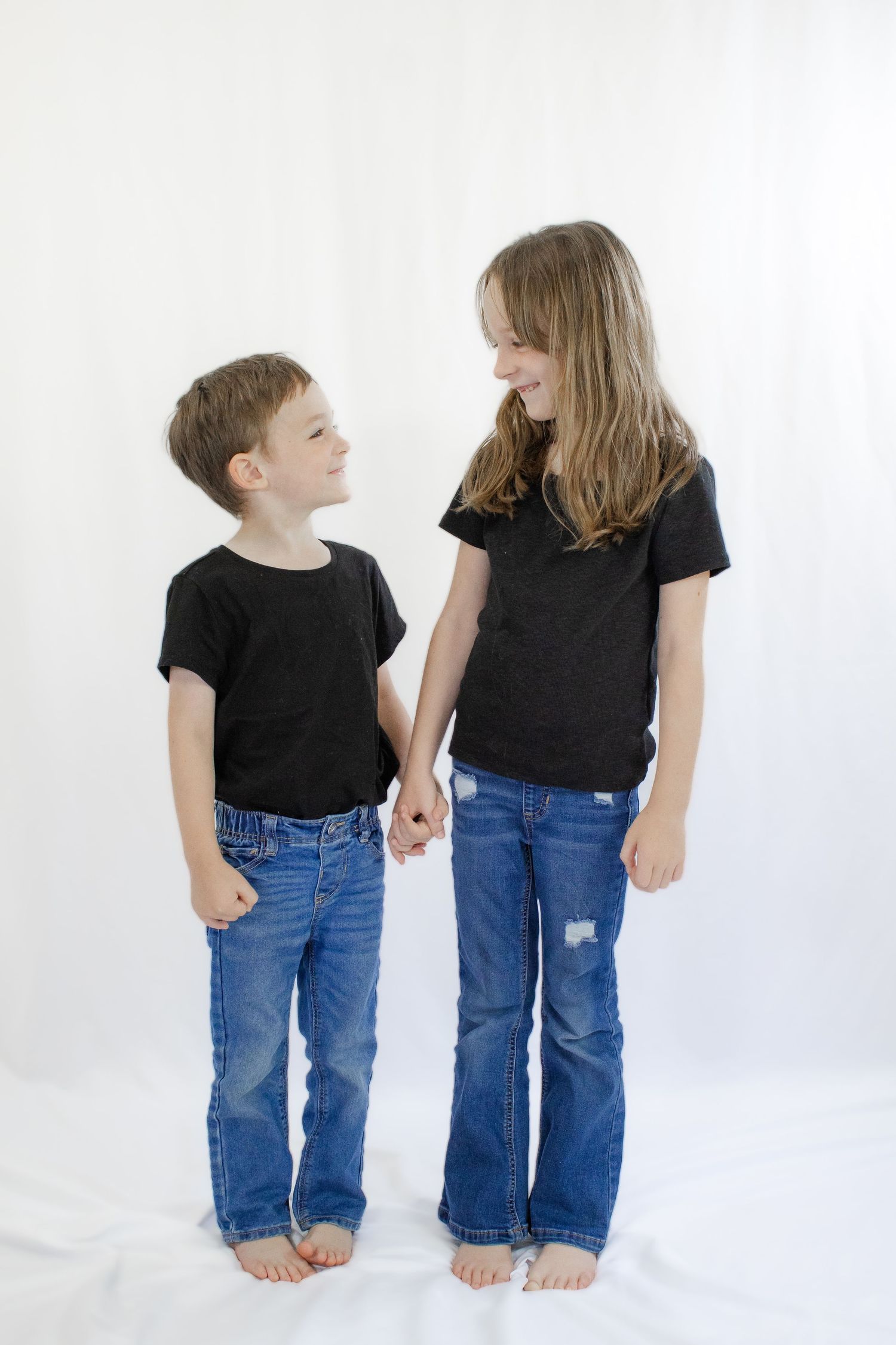 Two children in black shirts and jeans holding hands and smiling at each other against a white backdrop.