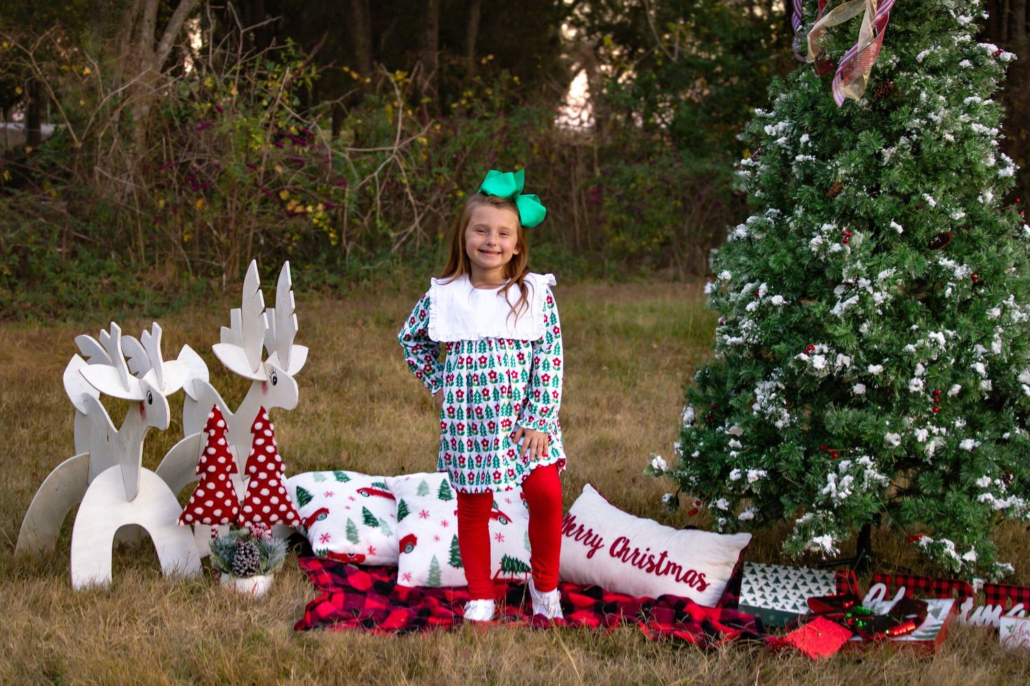 Girl in plaid dress and red leggings standing by Christmas tree with Merry Christmas pillow.