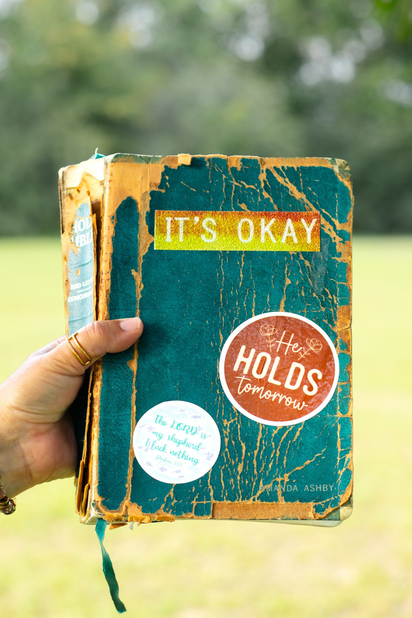 Hand holding worn teal Bible with stickers reading It's Okay and He Holds Tomorrow.
