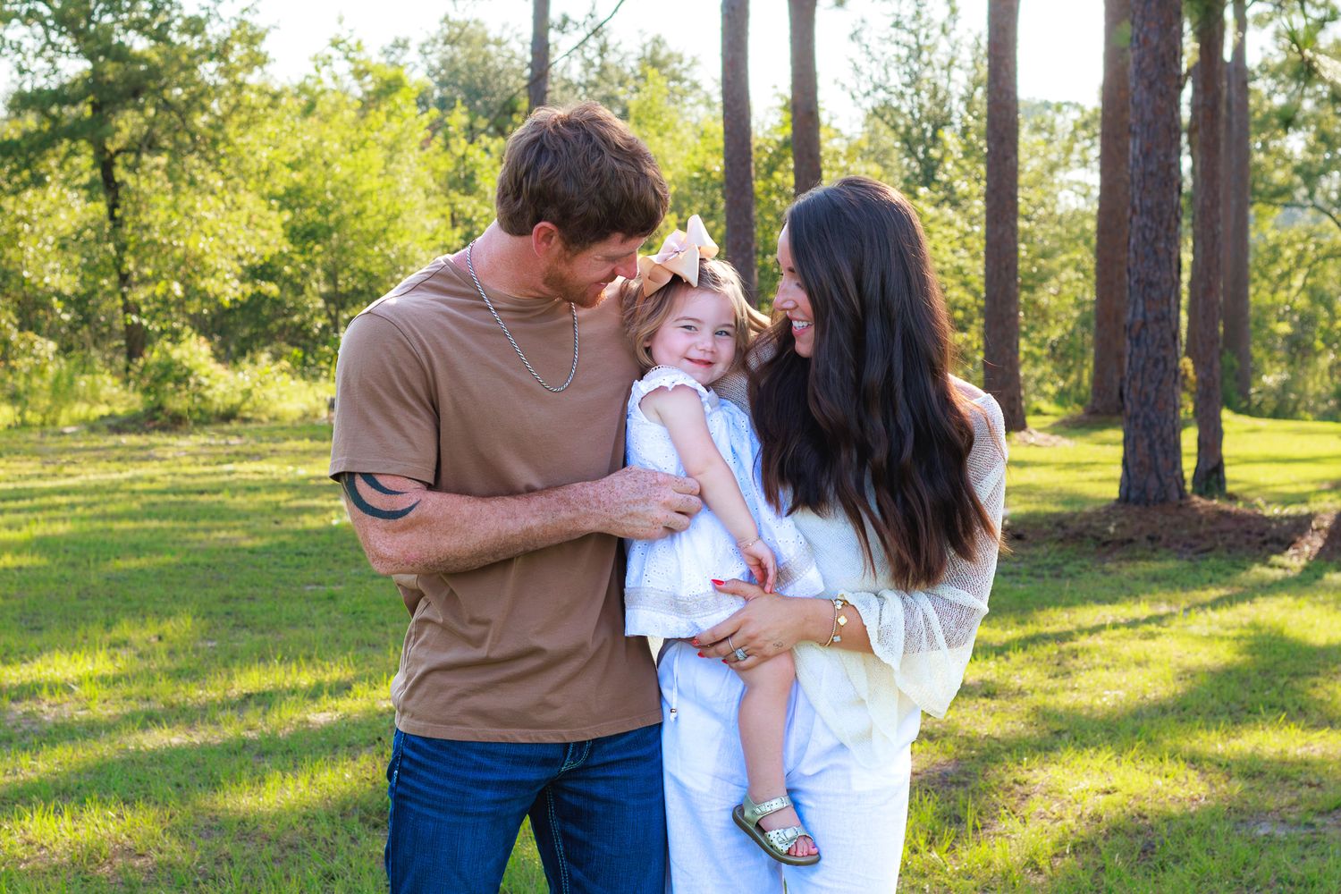 Young couple with long dark hair holds their toddler daughter in white dress among tall pine trees in sunny park.