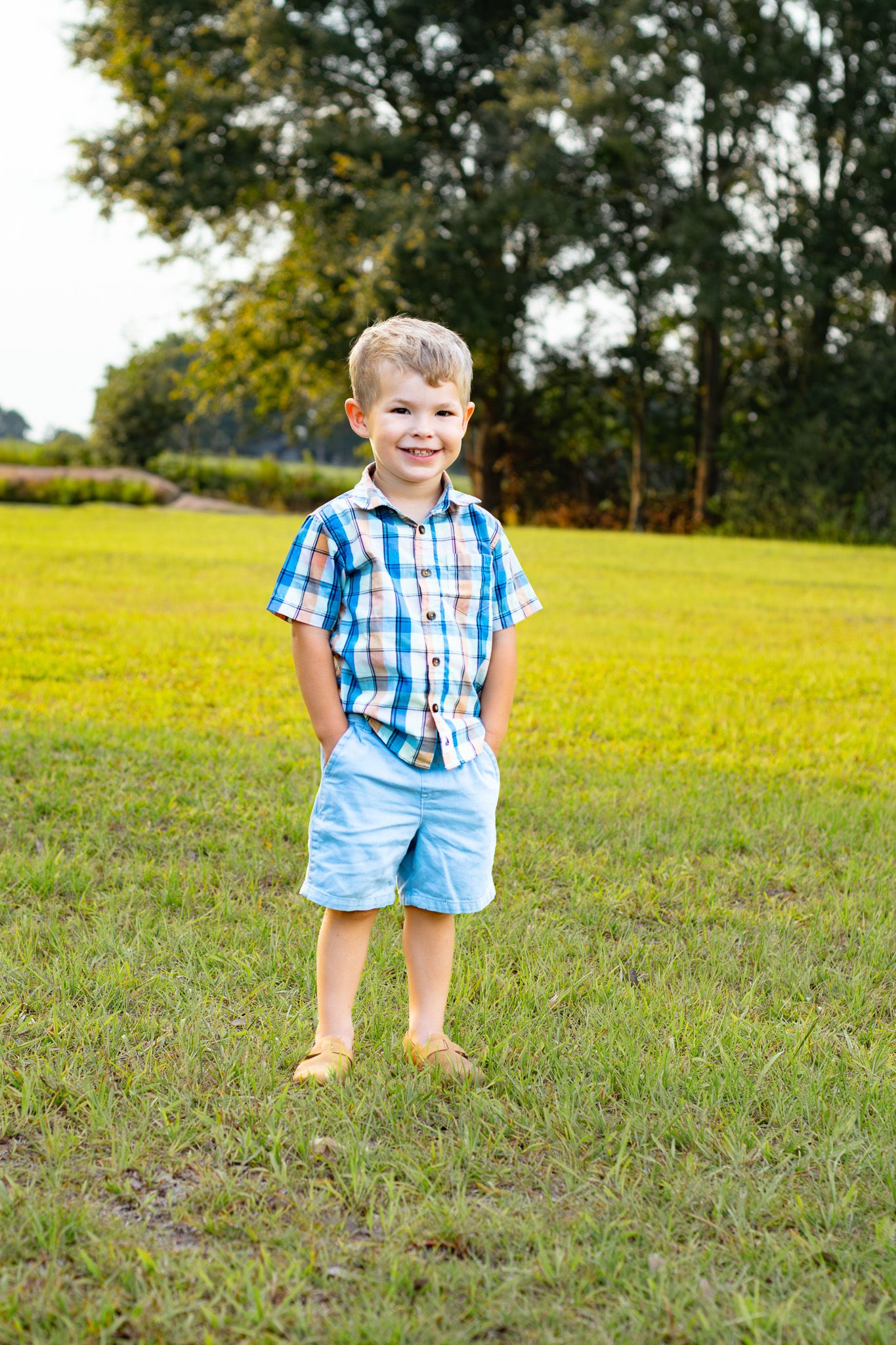 A smiling young boy in a plaid shirt and blue shorts stands on green grass with trees in the background.