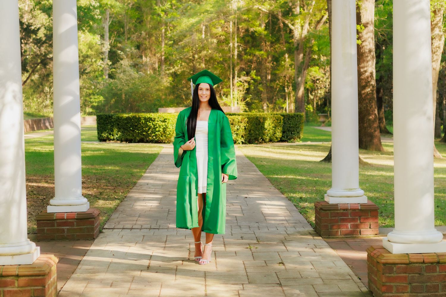 Young woman in green graduation cap and gown walking along brick pathway with white columns and lush greenery.