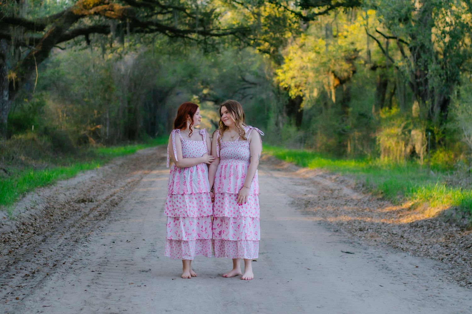 Two women in matching pink lace dresses stand face to face on a picturesque tree-lined dirt road.