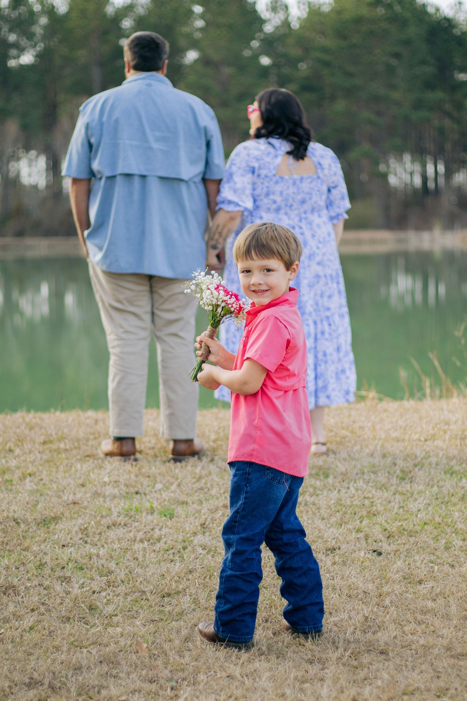 A smiling young boy in a pink shirt holds flowers while his parents stand by a pond.