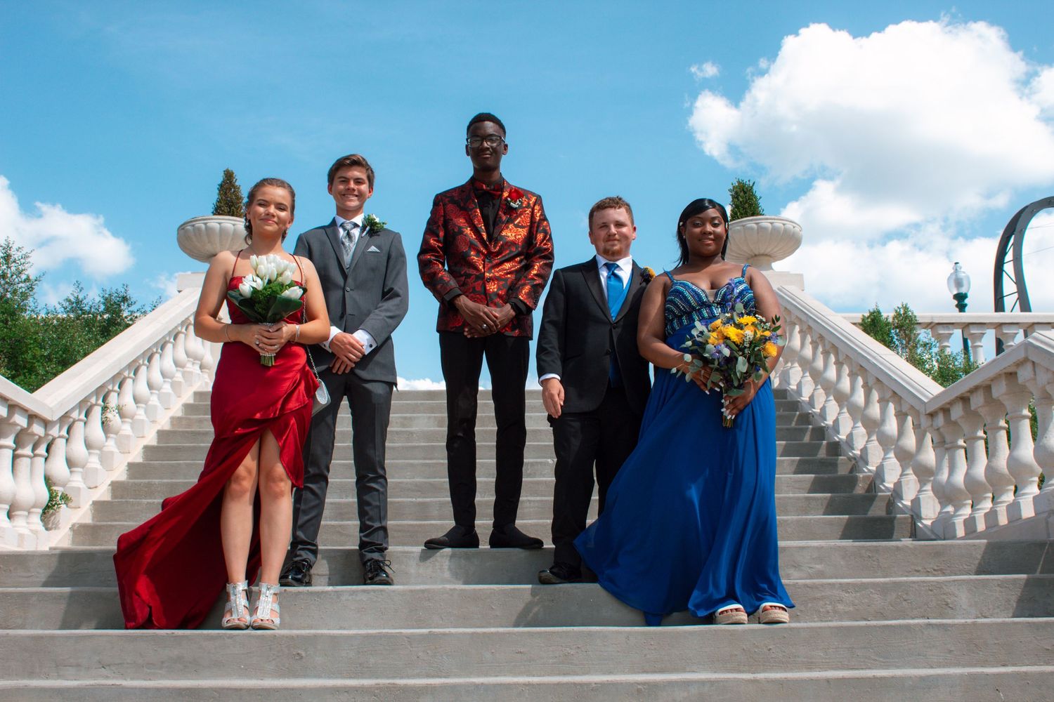 Five teenagers in formal prom attire posing on elegant white staircase under a bright blue sky.
