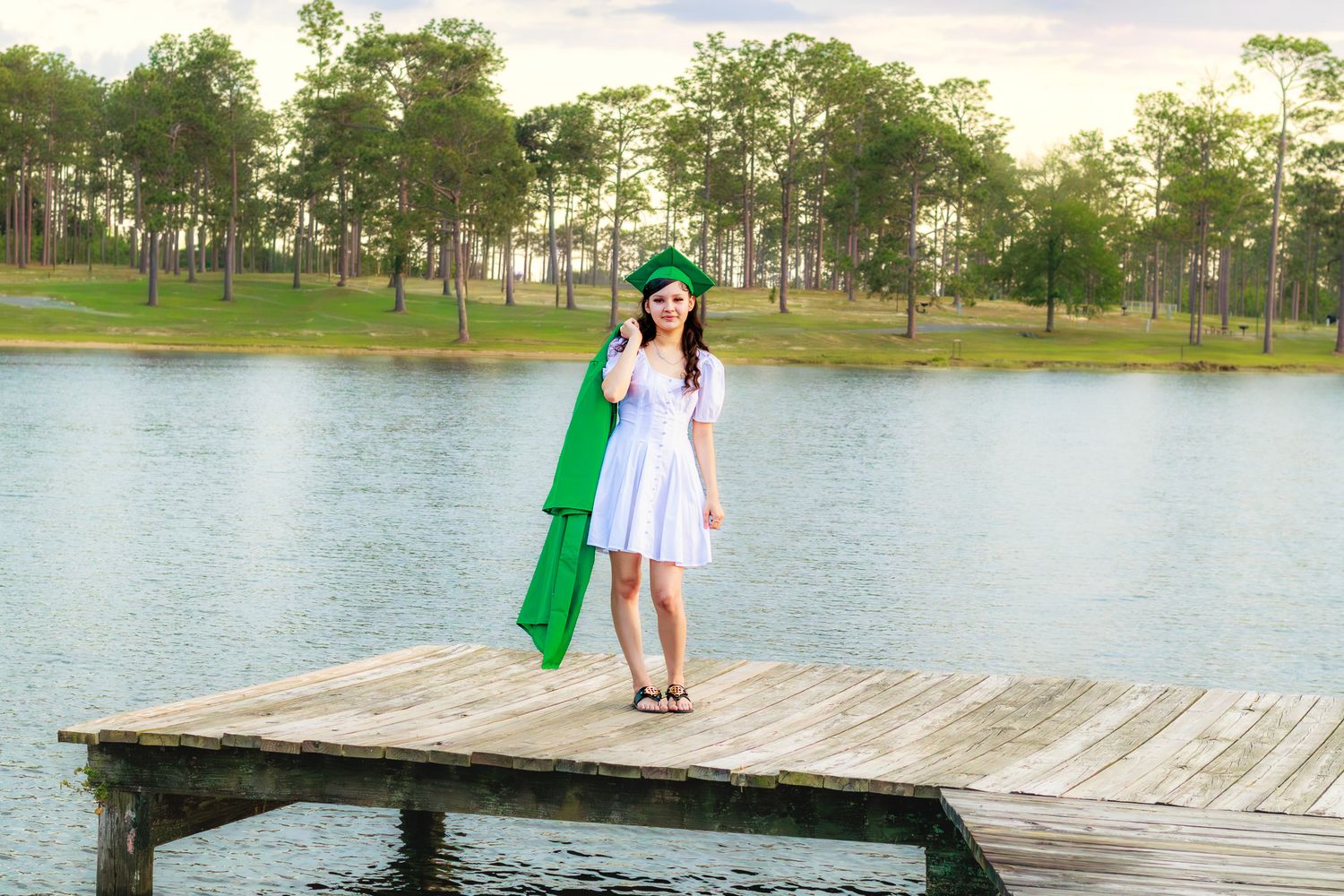 Graduate in white dress holding green cap and gown on wooden dock overlooking a serene lake.