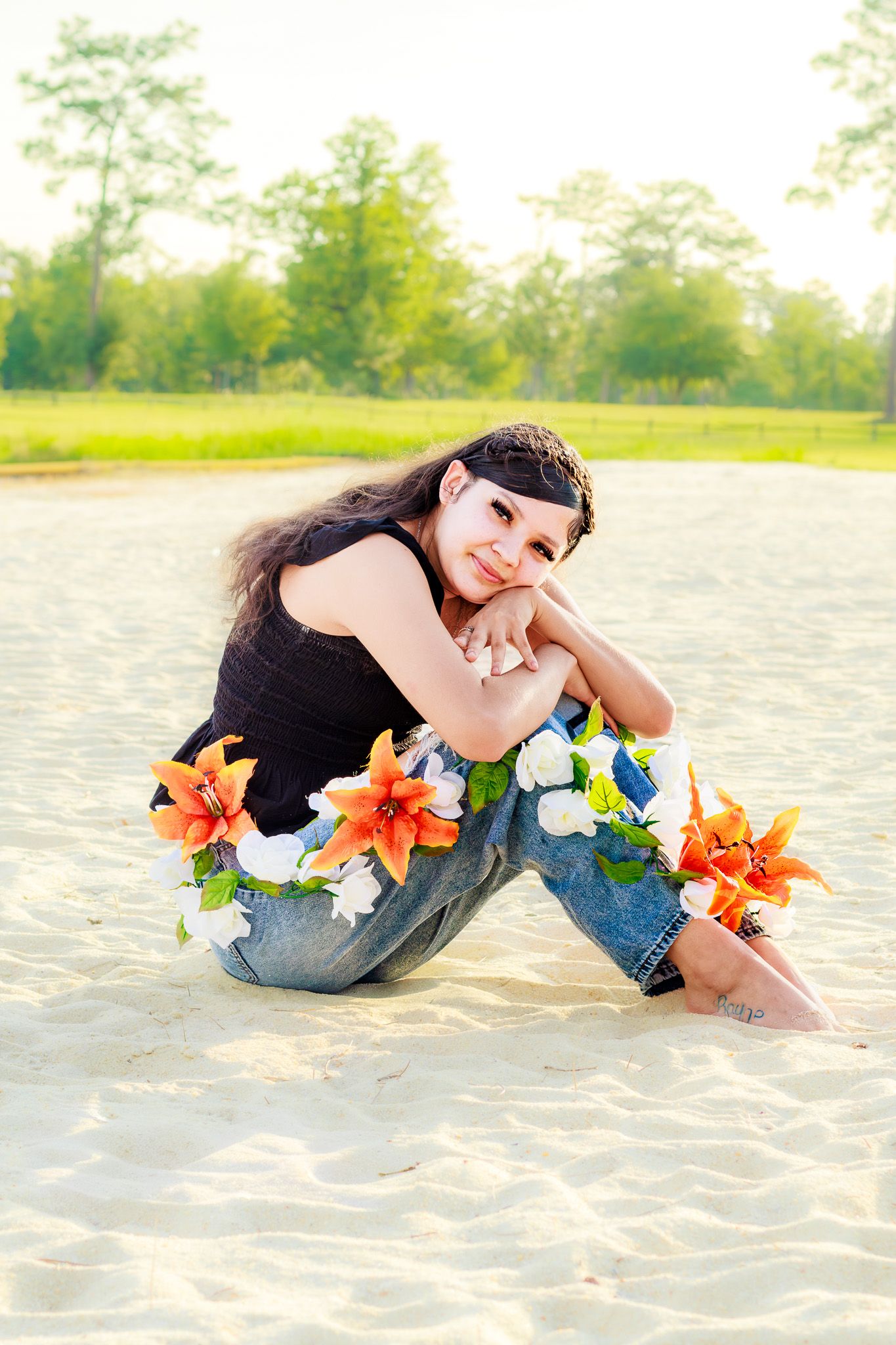 Young woman sitting on sandy beach wearing jeans adorned with colorful flowers, smiling warmly with green trees in background.