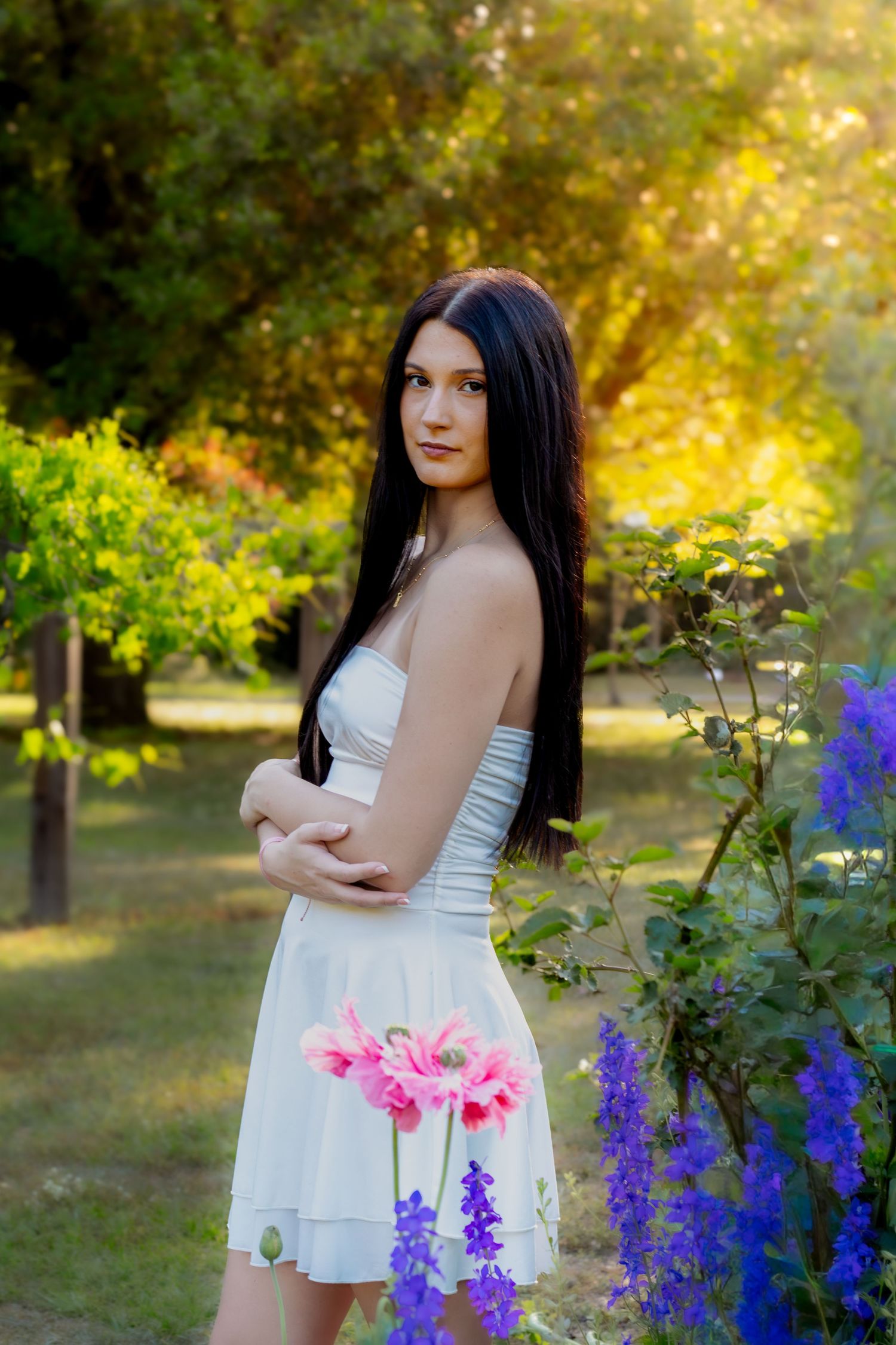 Young woman in white dress holding pink flowers in a sunlit garden with purple blooms and green trees.