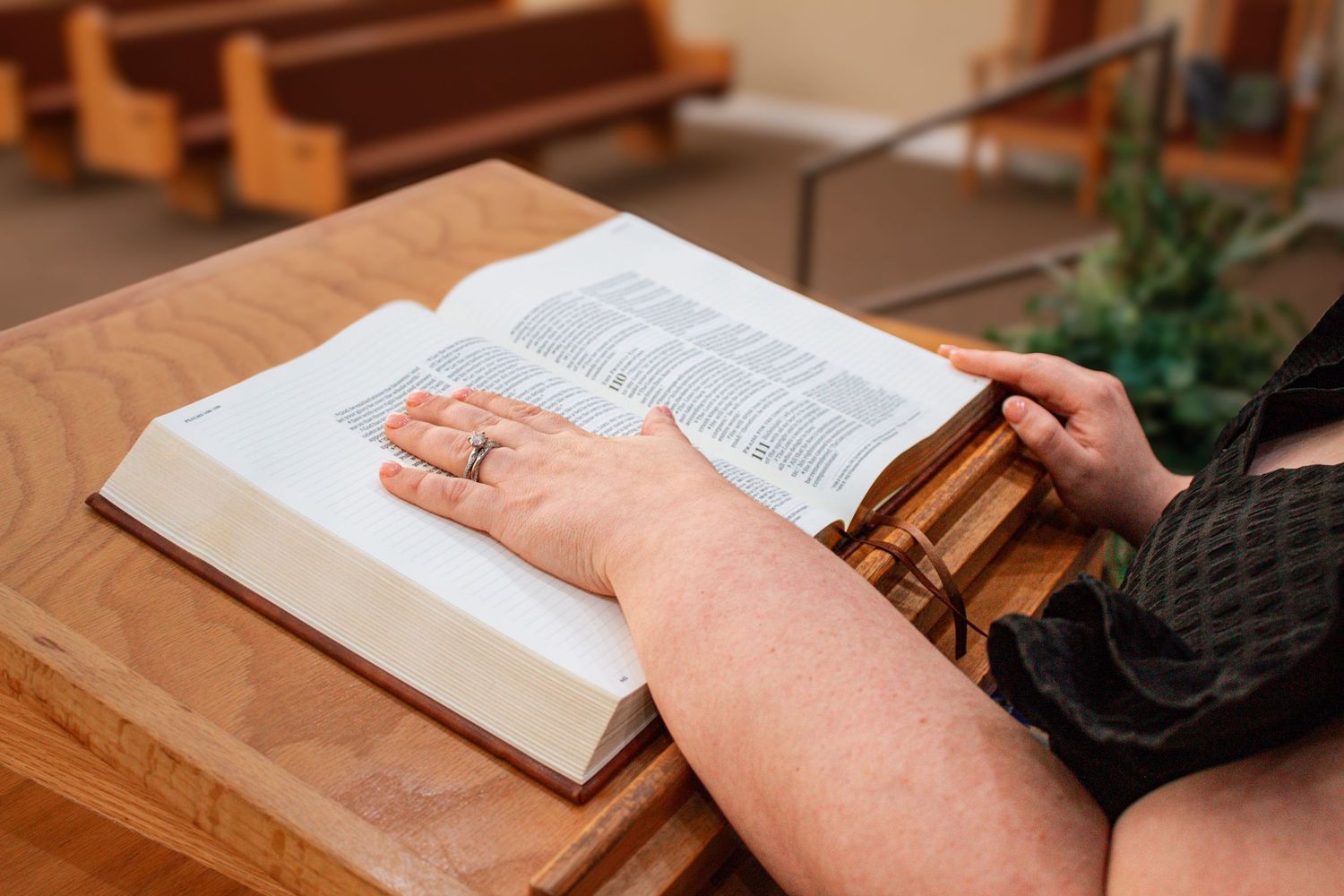 Woman's hand with ring resting on open Bible at wooden church lectern.
