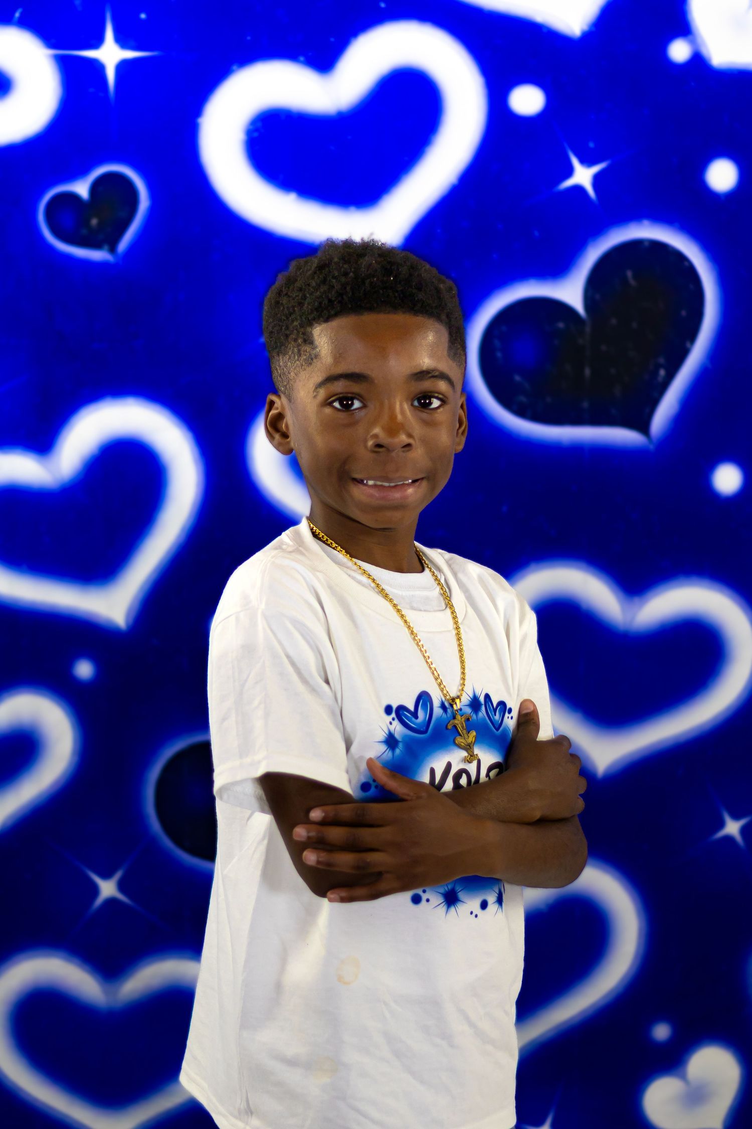 Smiling young Black boy in white tee with gold chain poses against blue hearts backdrop.