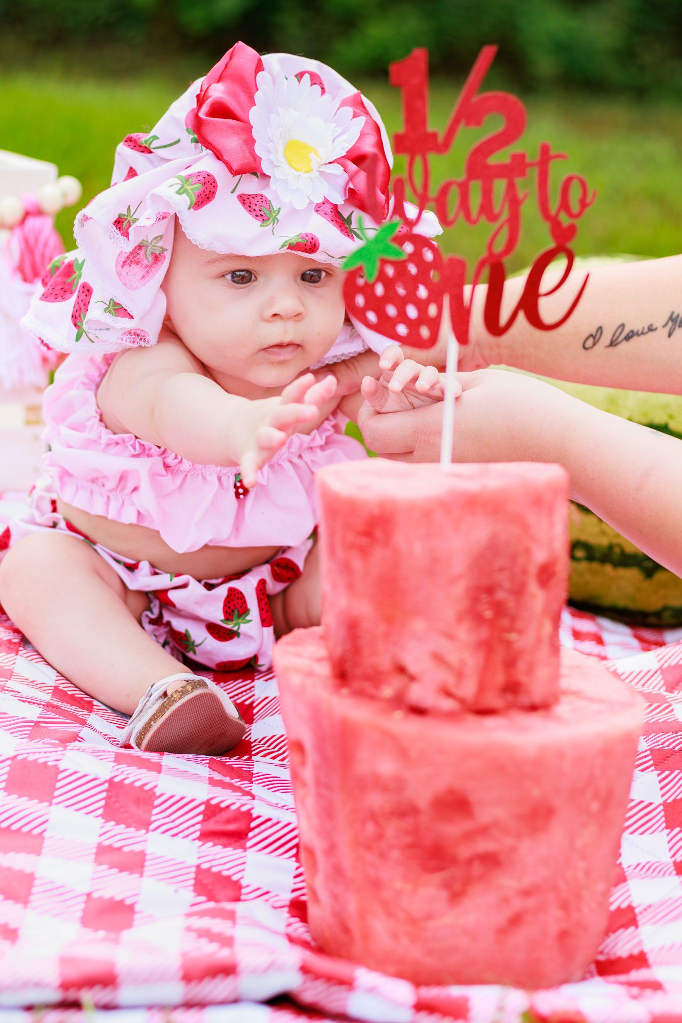 Baby girl in strawberry outfit celebrates half birthday with red cake and '1/2 Way to One' topper at outdoor picnic.
