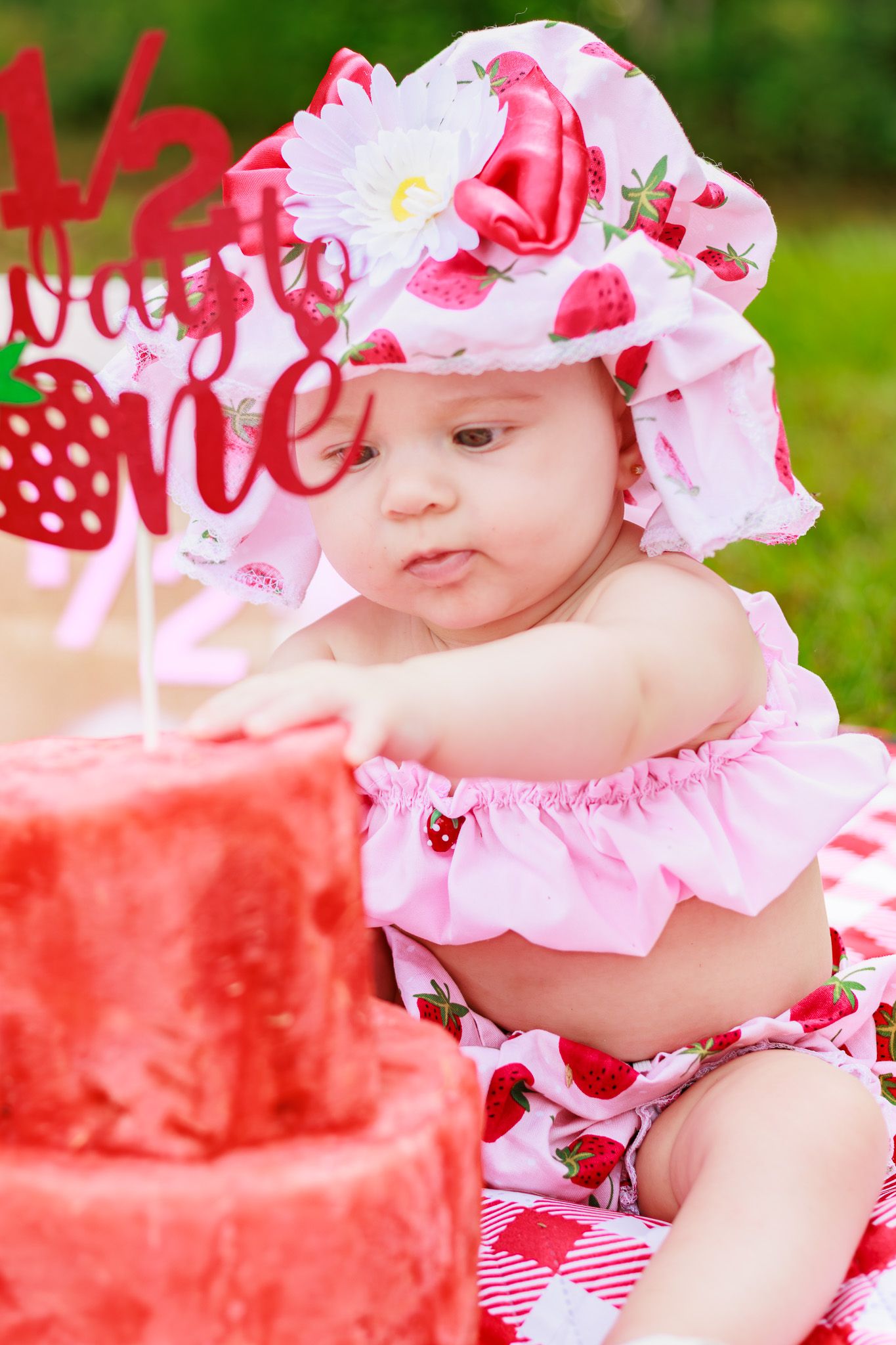 Baby girl in strawberry outfit and hat reaches for red smash cake at half birthday celebration outdoors.