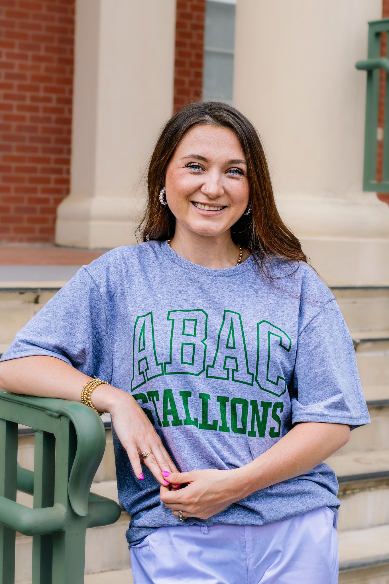 Smiling young woman in ABAC Stallions t-shirt leaning on green railing at brick building.