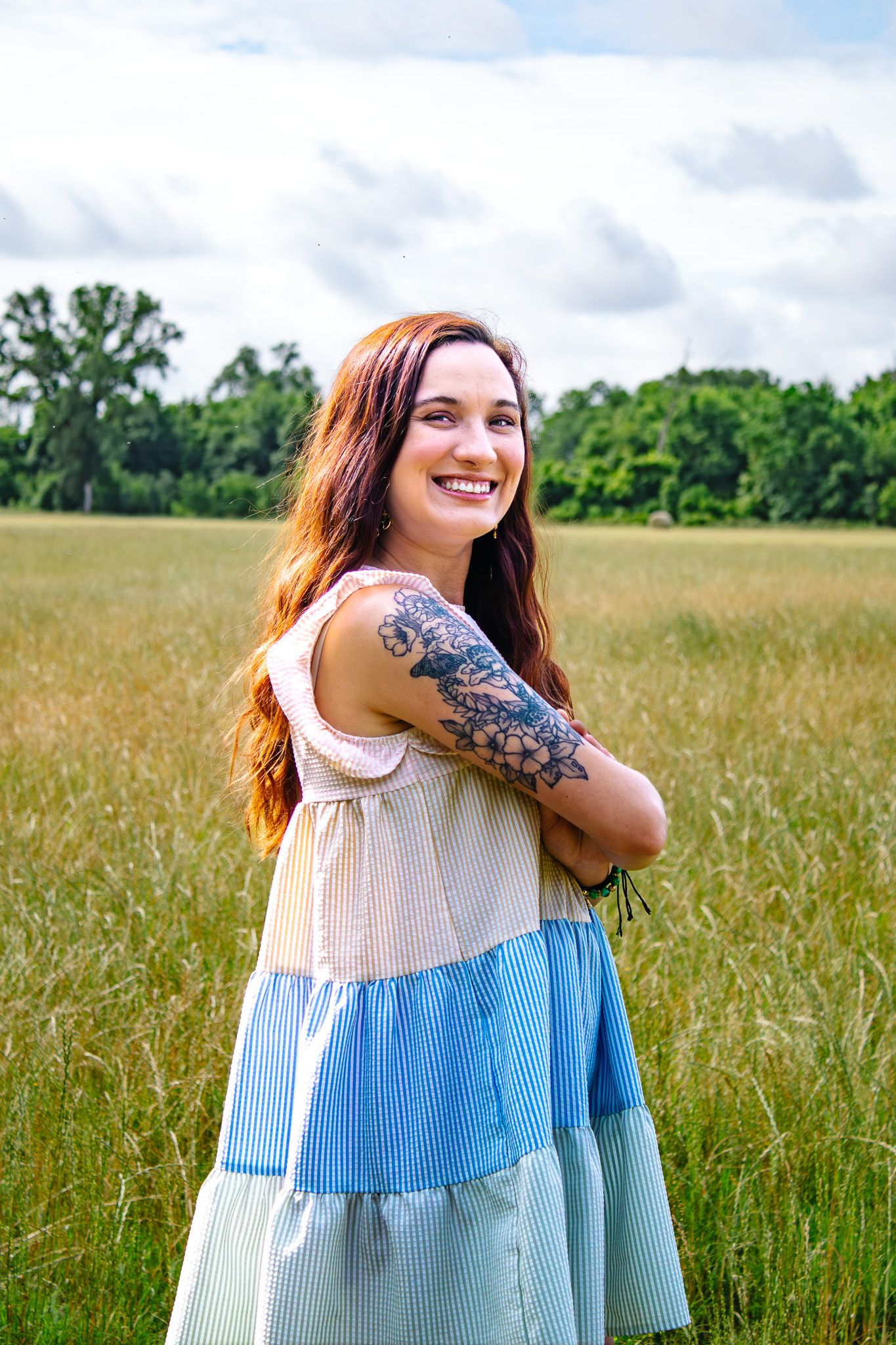 Smiling redhead woman with floral arm tattoo in a colorful tiered dress standing in a sunny grassy field.