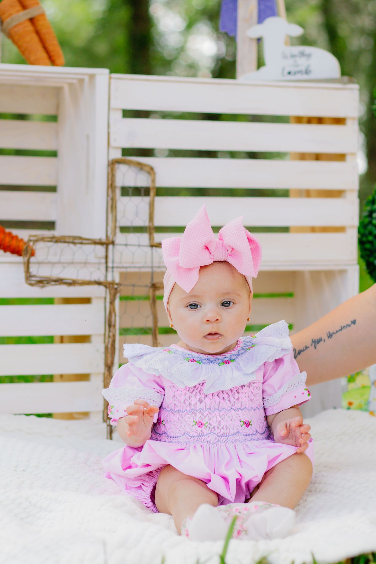 Baby girl in pink smocked dress with bow headband sitting near white wooden crates in garden setting.