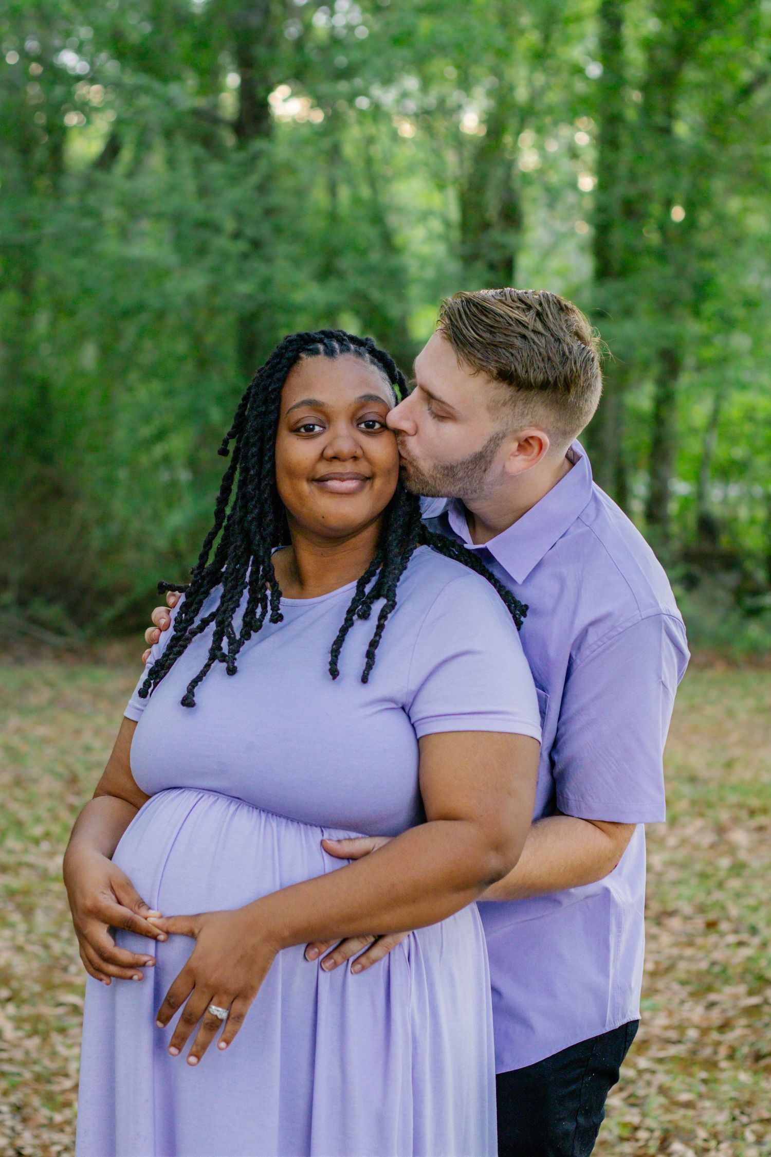 Pregnant woman in lavender dress smiles as partner kisses her cheek in forest setting.