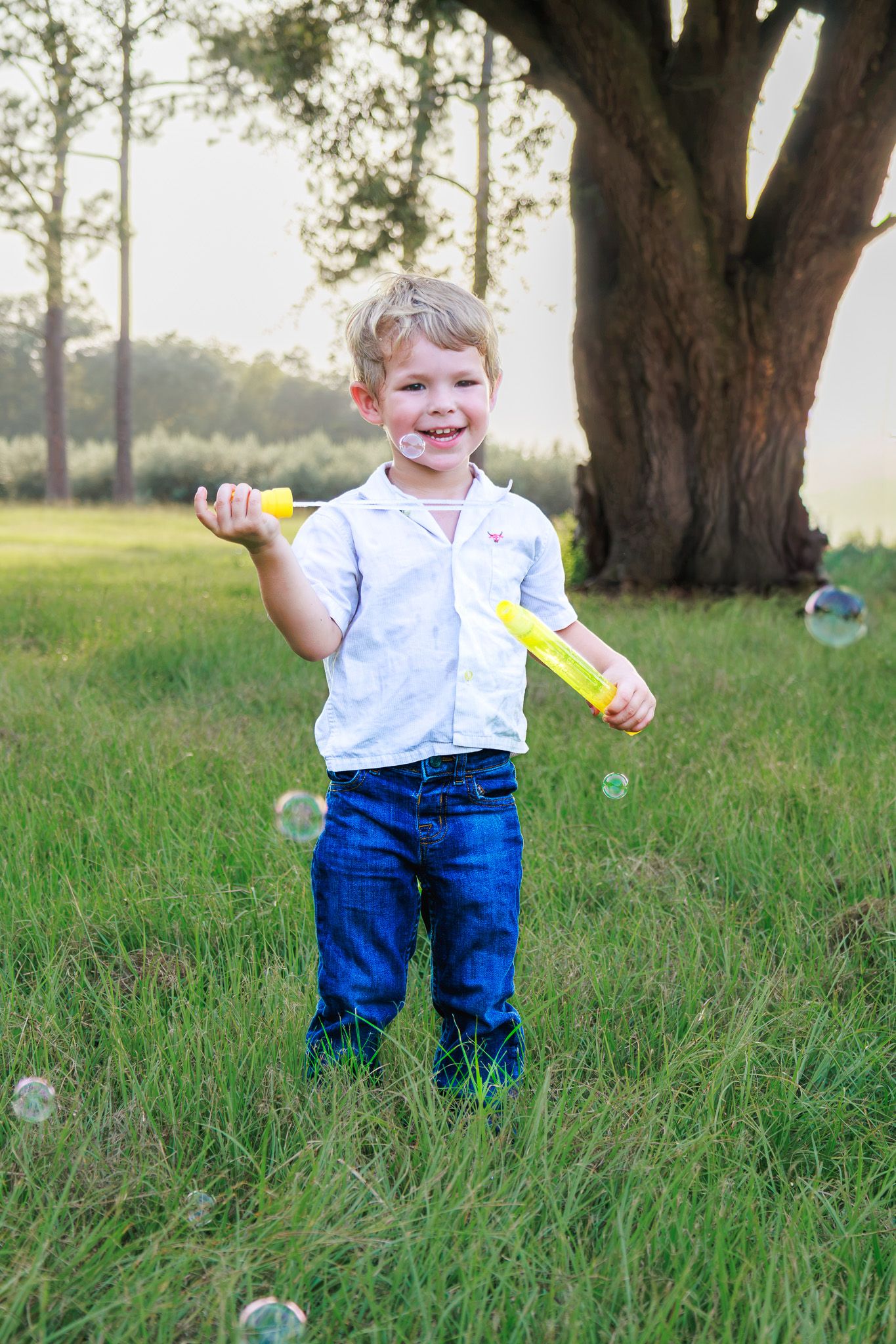 A happy young boy blowing bubbles outdoors in a green field, wearing a white shirt and blue jeans on a sunny day.