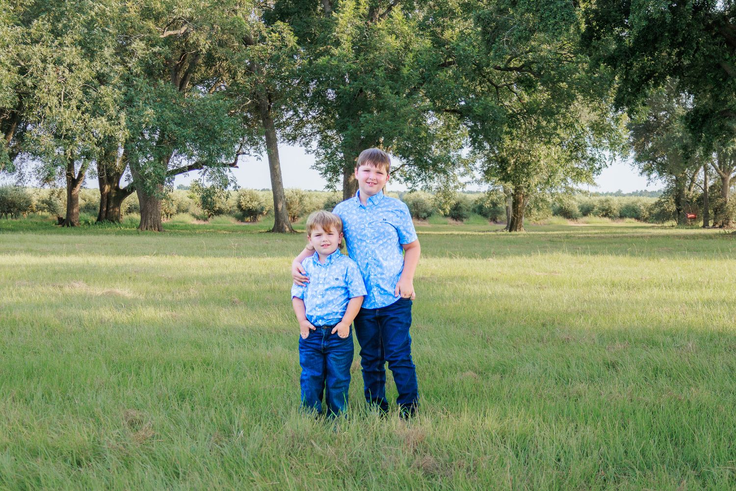 Two young boys in matching blue shirts and jeans pose together in a lush green field with trees in the background.