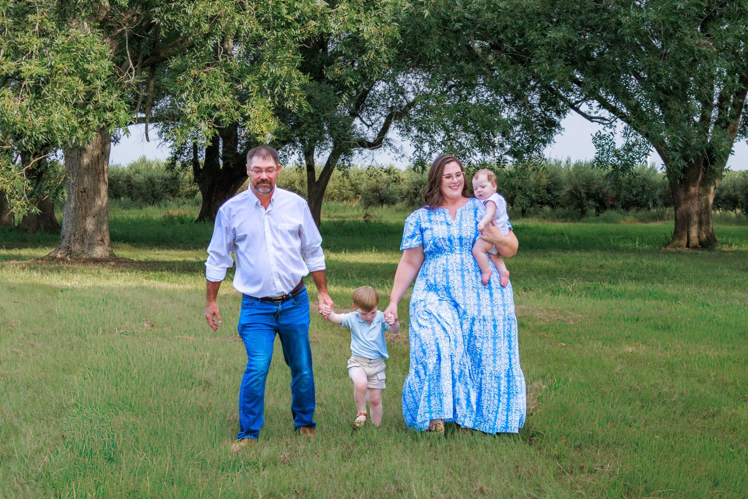 Family of four walking in green field, parents holding children's hands, wearing blue outfits, surrounded by trees.
